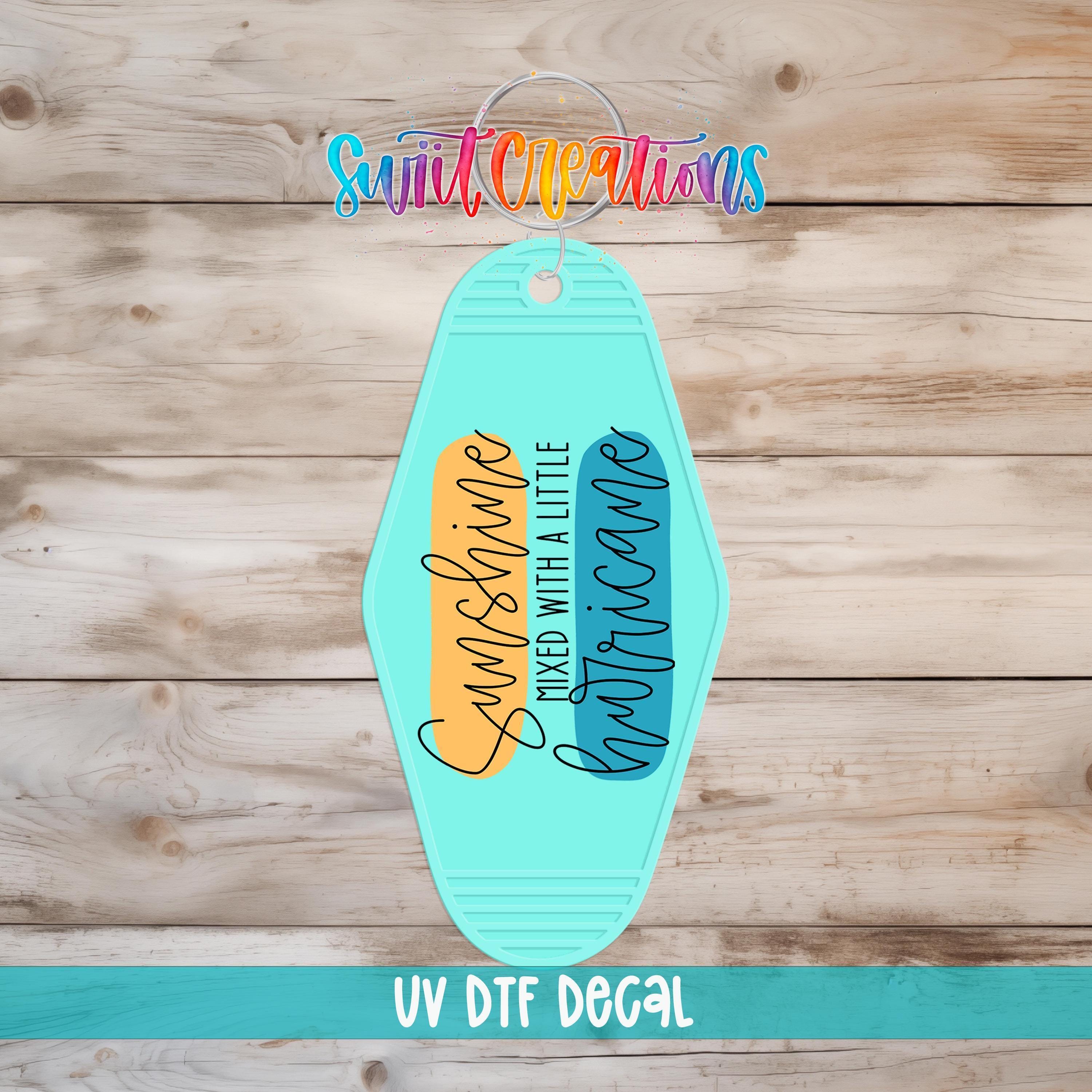 A UV DFT decal of a keychain with the text "Sunshine mixed with a little chaos" printed on it, set against a wooden background.