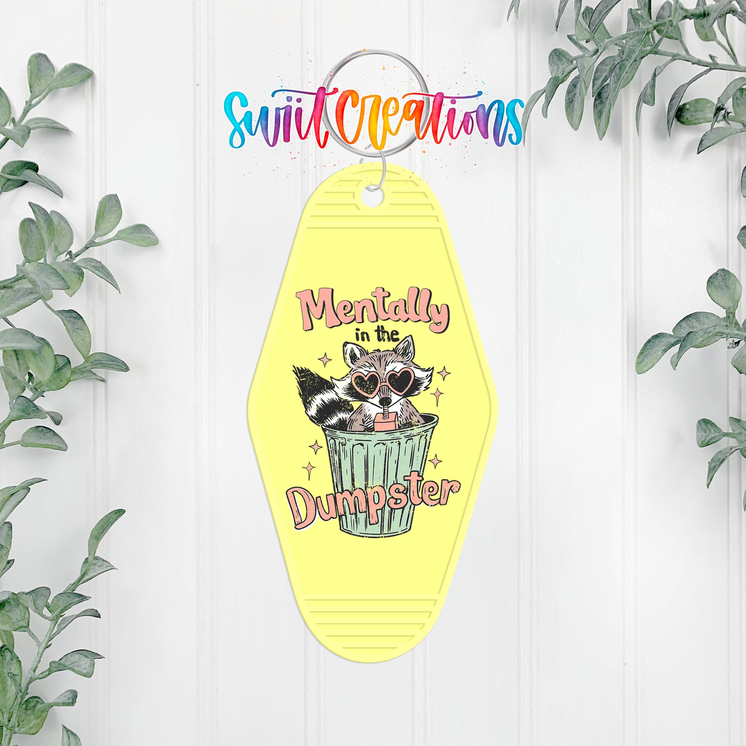 A yellow keychain with a cartoon raccoon sitting in a trash can and the text "Mentally in the Dumpster" printed on it.