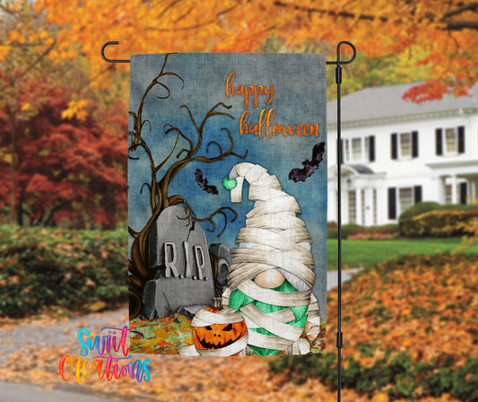 A Halloween-themed garden decoration featuring a gnome wrapped in bandages, a carved jack-o-lantern, and a gravestone with the word "RIP" on it, set against a backdrop of autumn foliage.