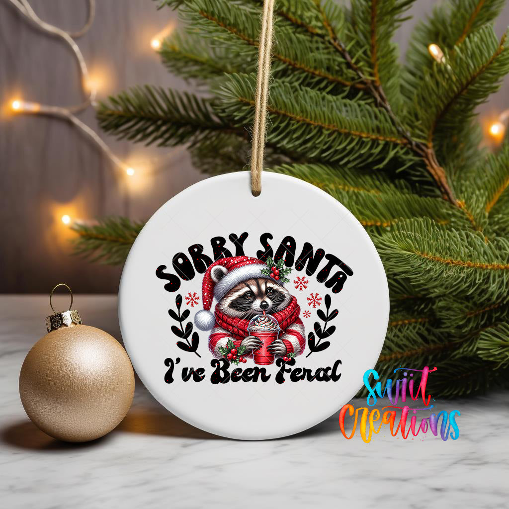 A white ceramic ornament with a raccoon wearing a Santa hat and scarf, holding a mug, and the text "Sorry Santa, I've been fired" is displayed on a table next to a gold ornament and a Christmas tree.