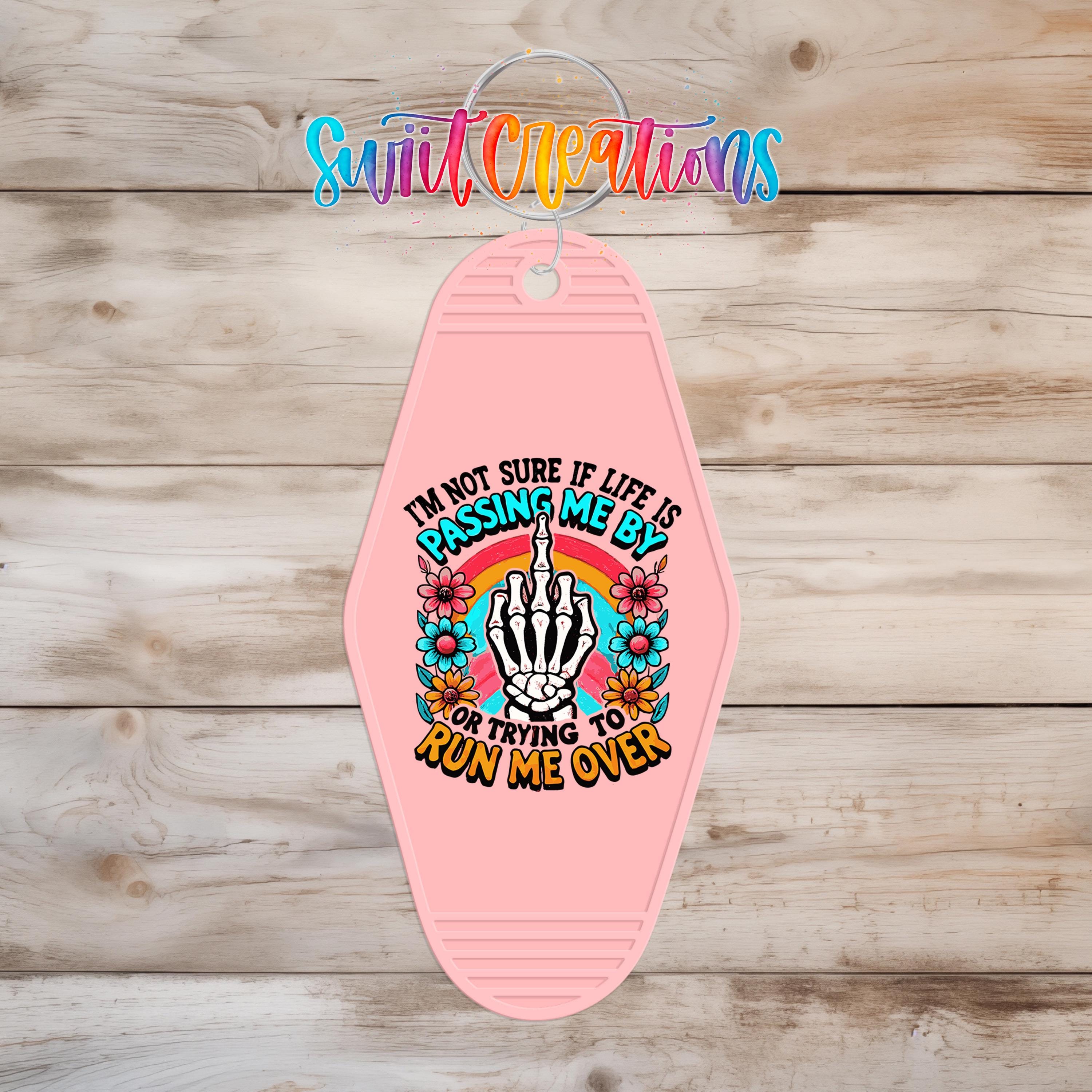A pink keychain with a graphic design featuring a skeleton hand and the text "I'm not sure if life is passing me by or trying to run me over".