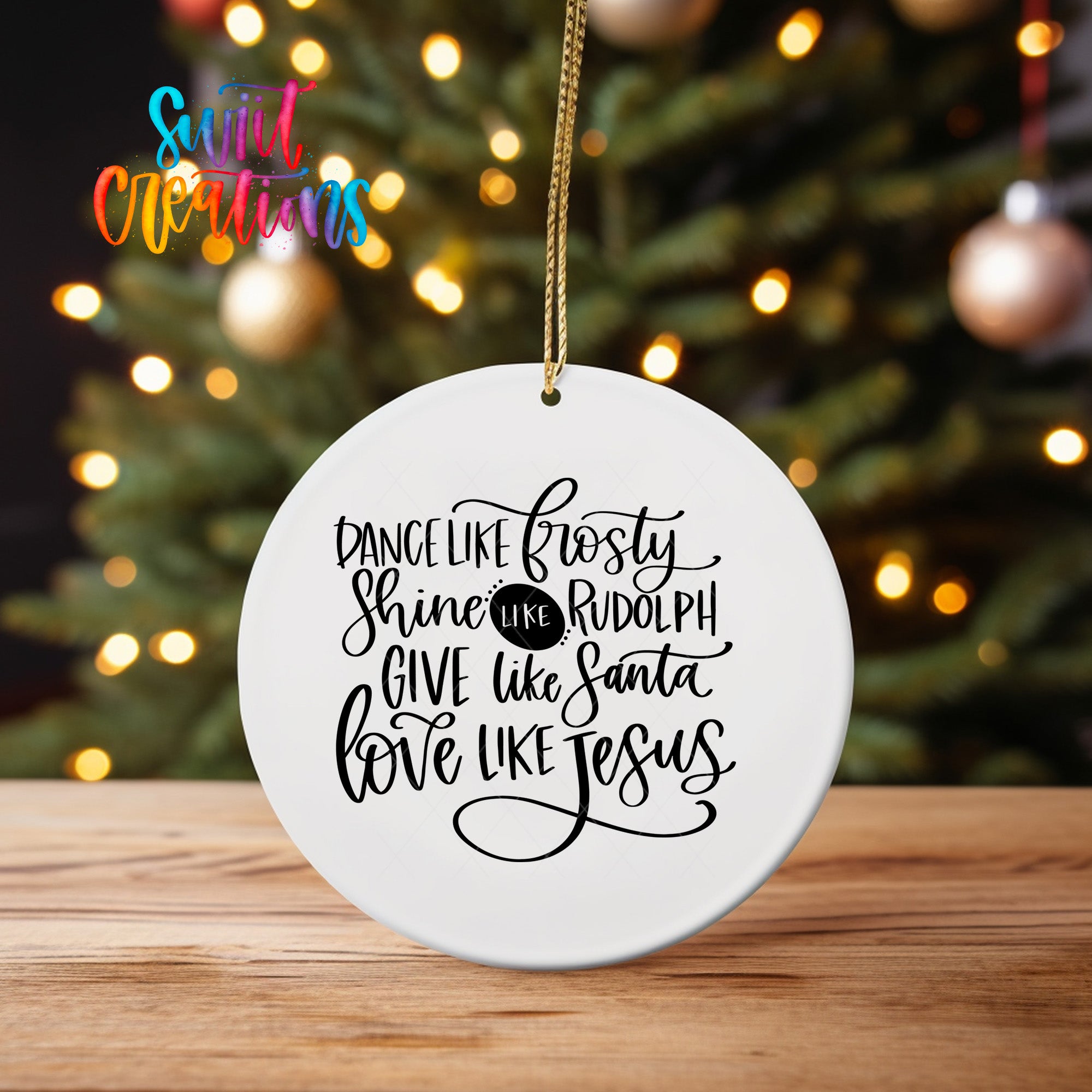 A white ceramic ornament with a black text design hangs in front of a blurred Christmas tree with colorful lights.
