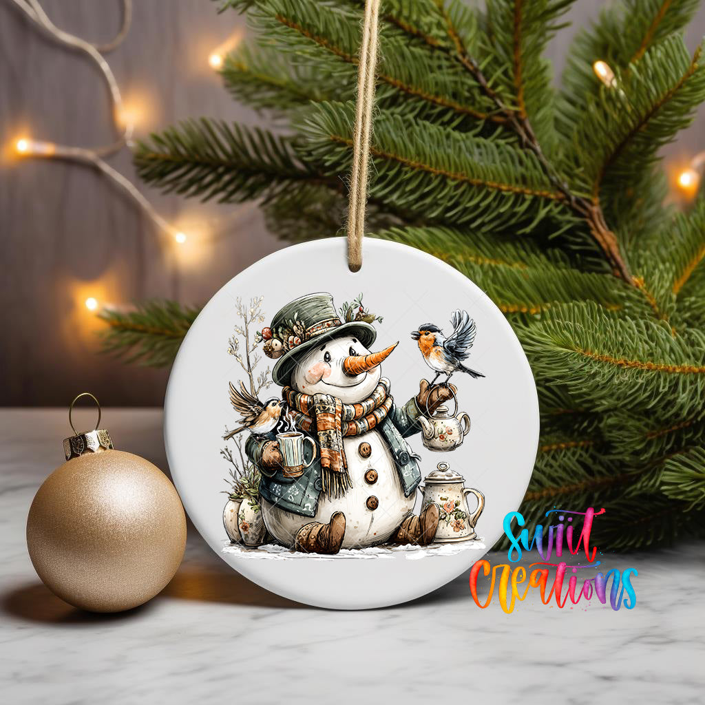 A white ceramic ornament featuring a snowman holding a bird and a teapot, surrounded by pine branches and a Christmas tree with twinkling lights in the background.