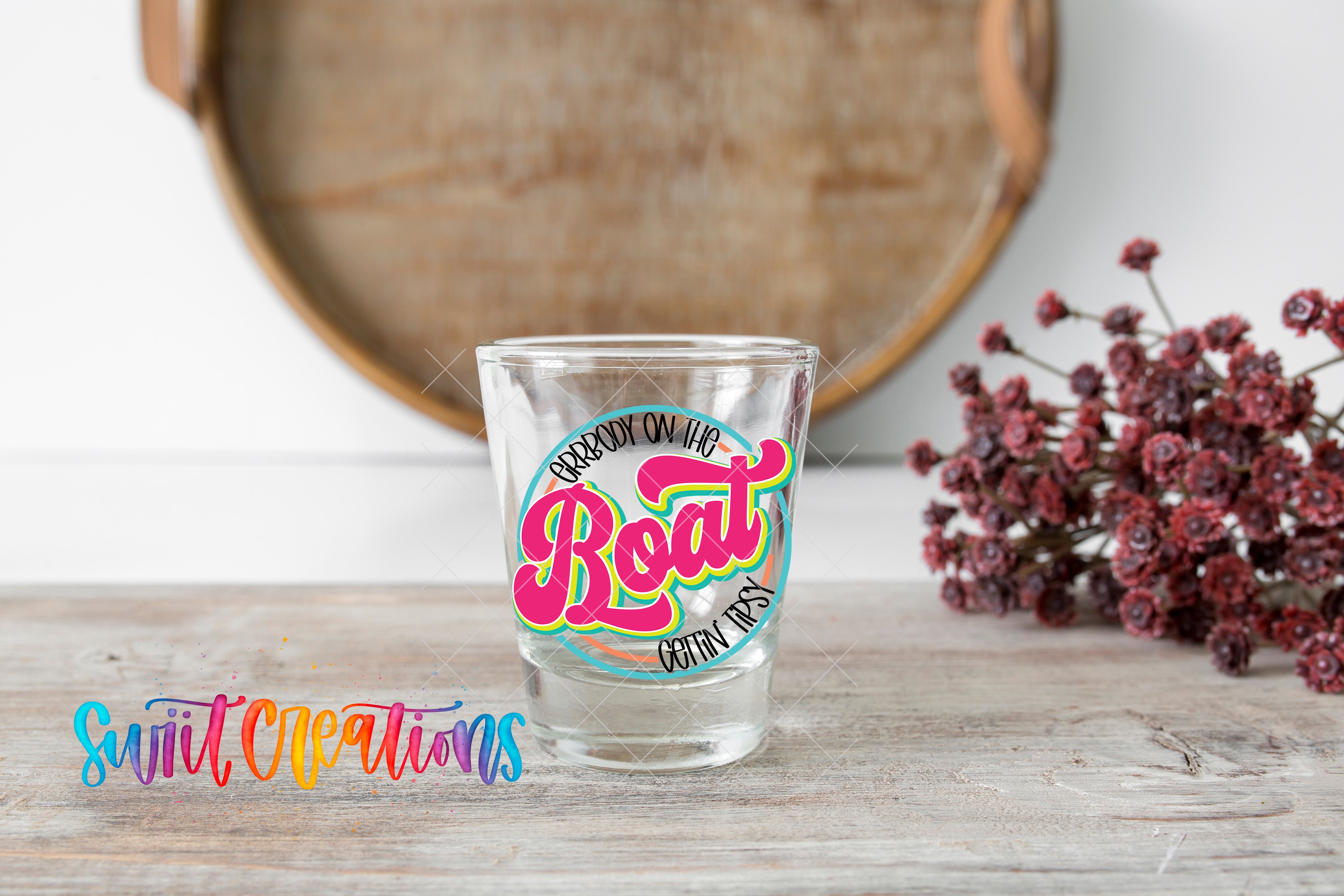 A shot glass with a colorful label that says "Boogie Boogie Boogie" is placed on a wooden surface, with a bunch of red flowers in the background.