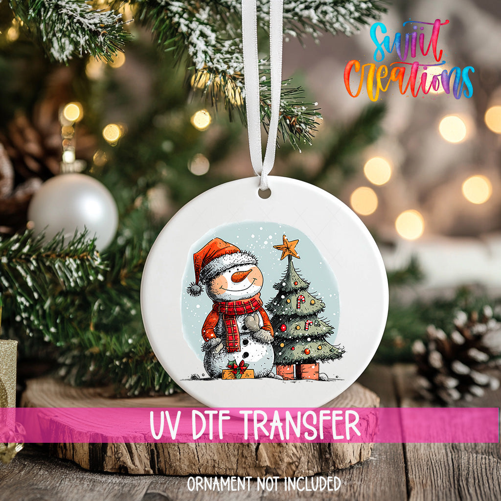 A white ornament with a snowman and Christmas tree design hangs on a Christmas tree.