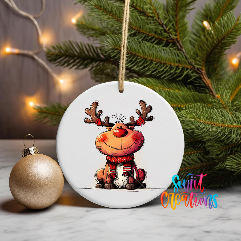A white ceramic ornament featuring a cartoon reindeer with antlers, sitting on a white surface next to a gold ornament and a Christmas tree with twinkling lights.