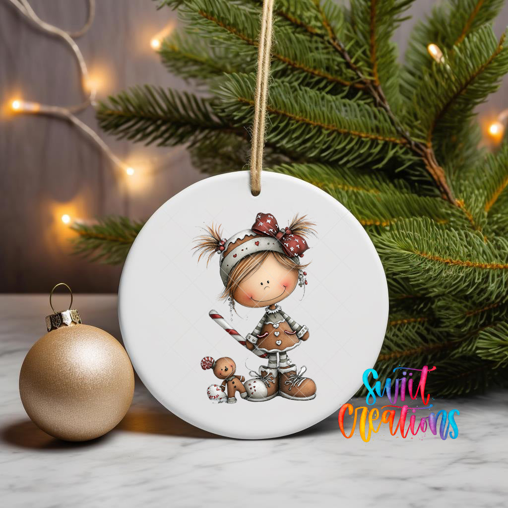 A white ceramic ornament featuring a smiling girl with a gingerbread man companion, placed on a marble surface next to a gold ornament and a Christmas tree with twinkling lights.