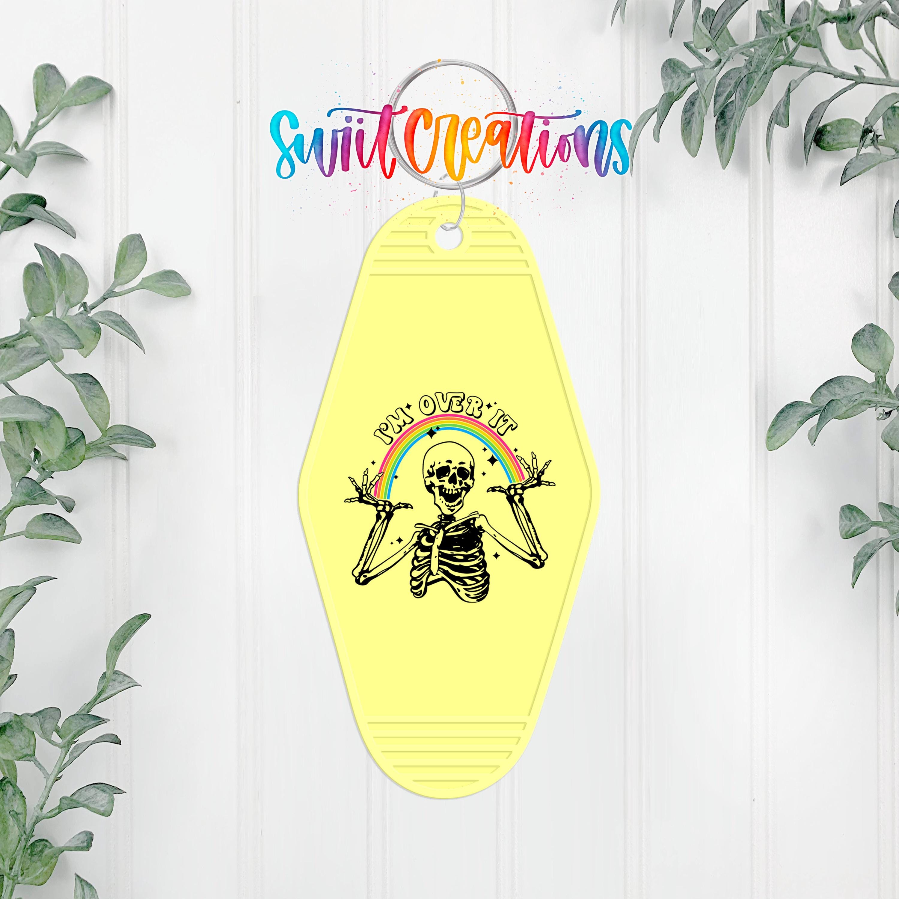 A yellow keychain with a skeleton graphic and a rainbow graphic, hanging on a white wall with greenery in the background.