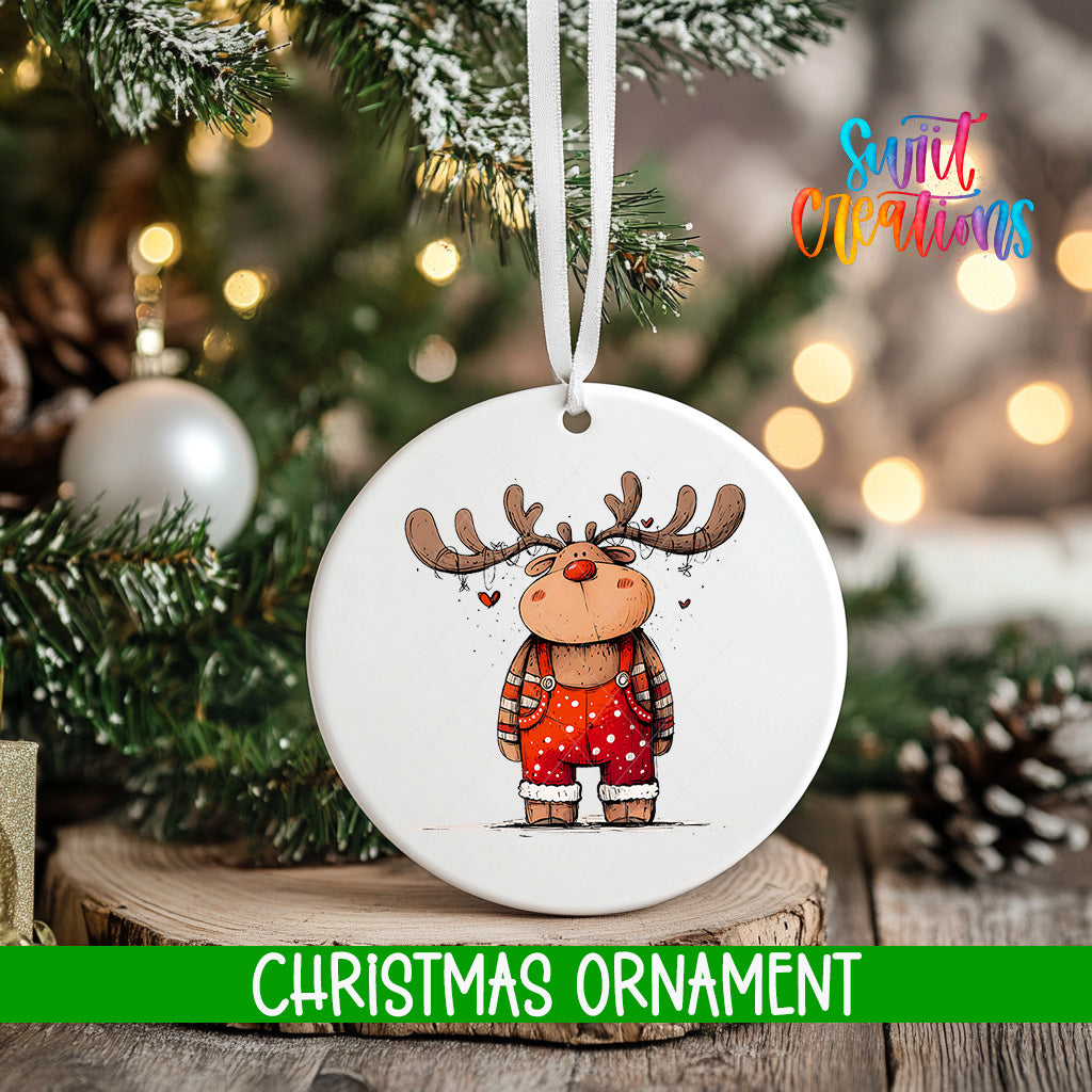 A white Christmas ornament featuring a cartoon reindeer with antlers and overalls, hanging on a tree branch with a blurred background of lights and greenery.
