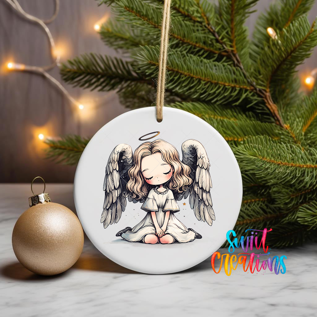 A white ceramic ornament with a cartoon illustration of a young girl with angel wings, sitting on her knees, is displayed on a table next to a gold ornament.