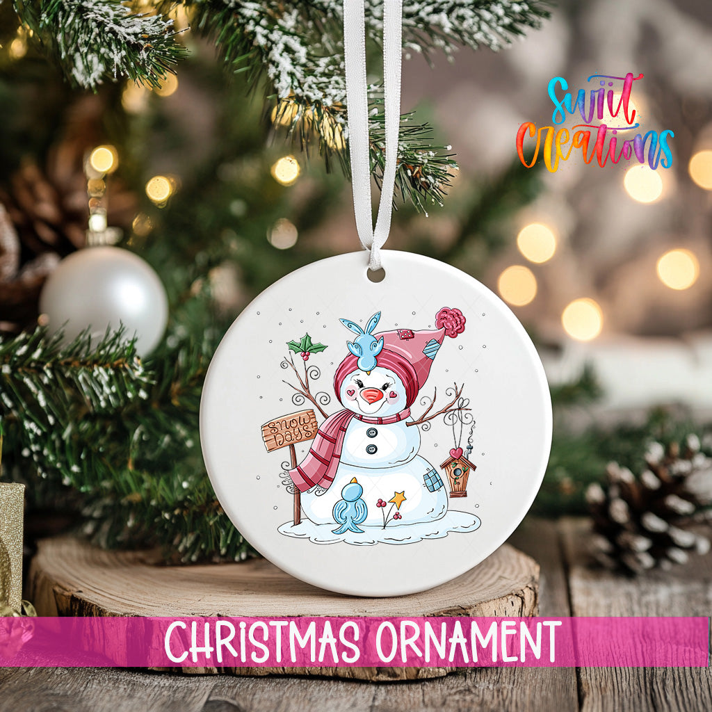 A white Christmas ornament featuring a snowman and other winter-themed elements is hanging from a tree branch.