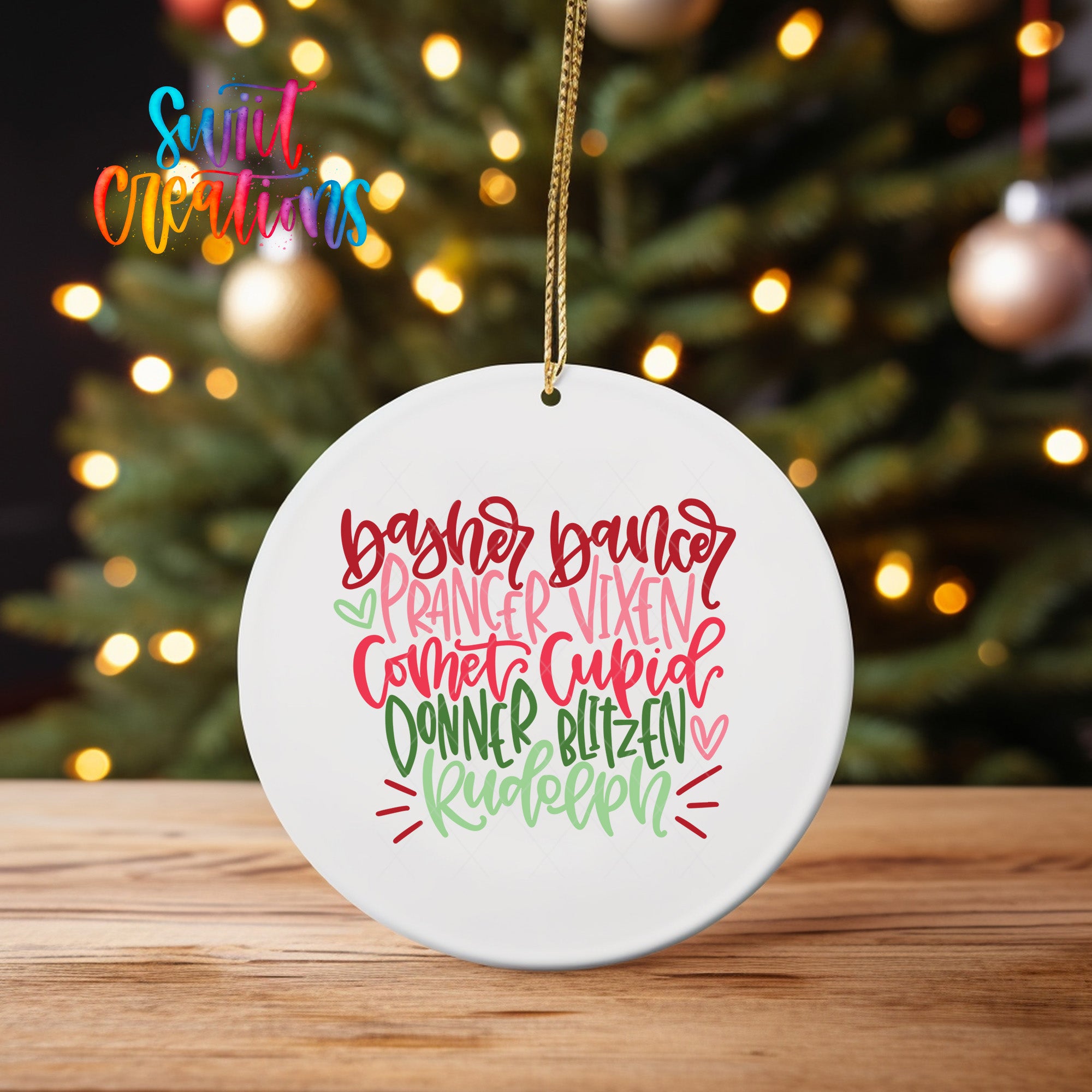 A white ceramic ornament with a festive design hangs on a wooden surface, with a blurred background of a Christmas tree and twinkling lights.