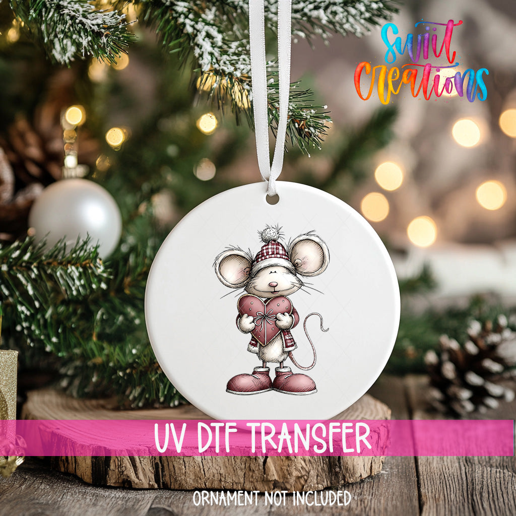 A white ornament with a mouse wearing a red hat and holding a heart, hanging on a Christmas tree.