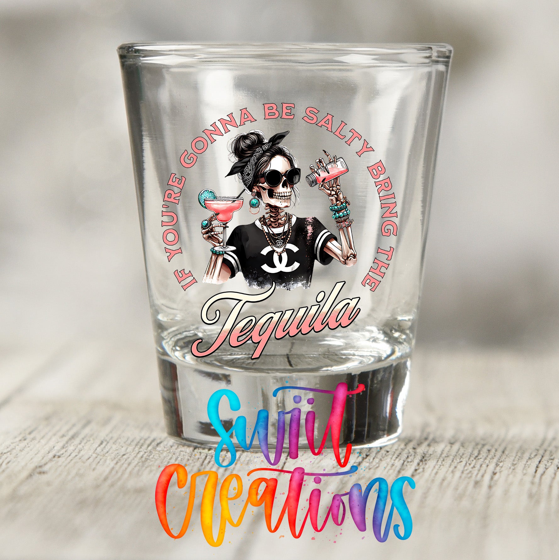 A shot glass with a graphic design featuring a skeleton wearing sunglasses and holding a tequila drink, along with the text "If you're gonna be salty bring the tequila" and the word "Sujiv creations" written below.