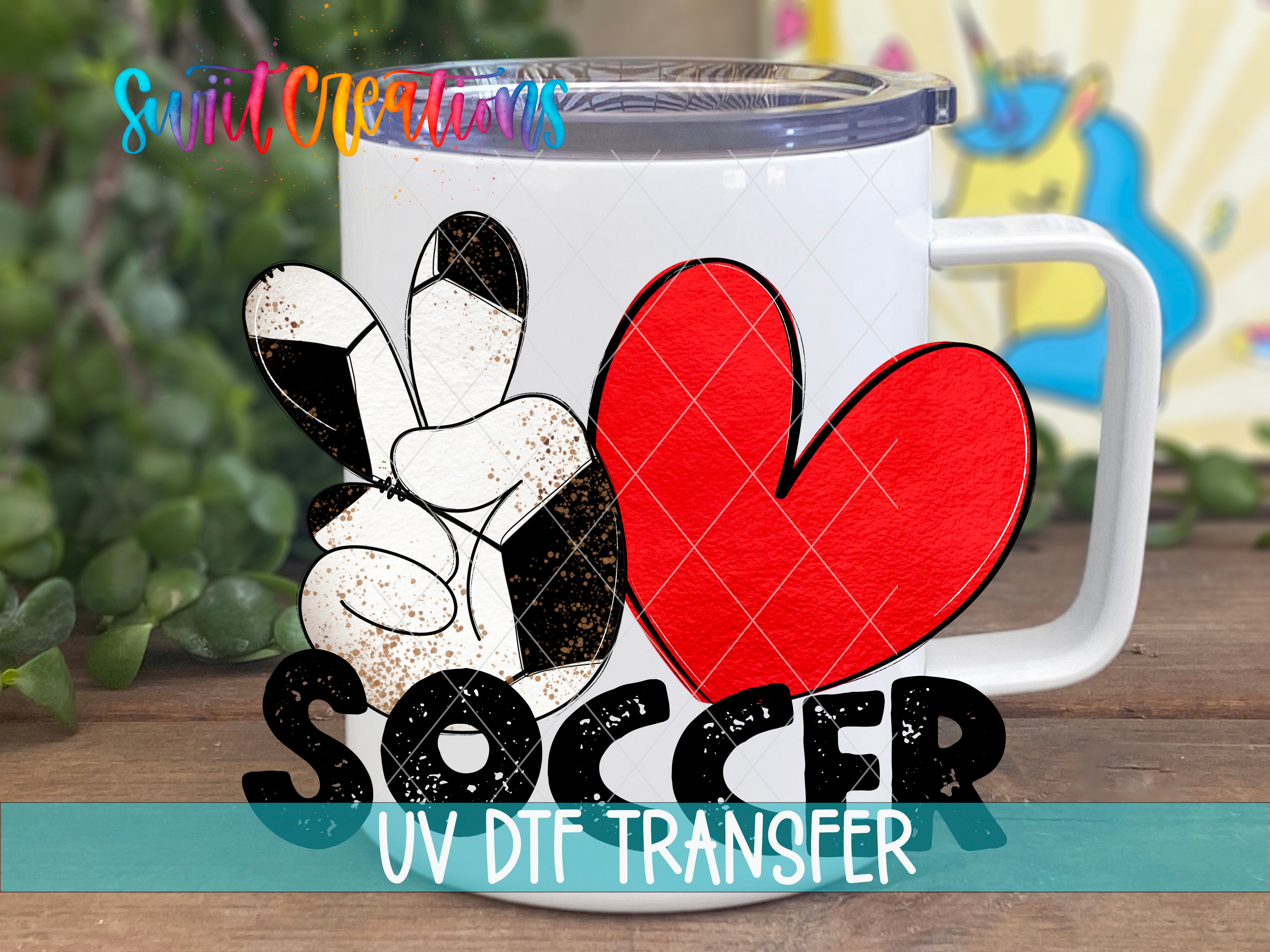 A white mug with a peace sign and a red heart, along with the word "SOCCER" printed on it, is displayed on a wooden surface.