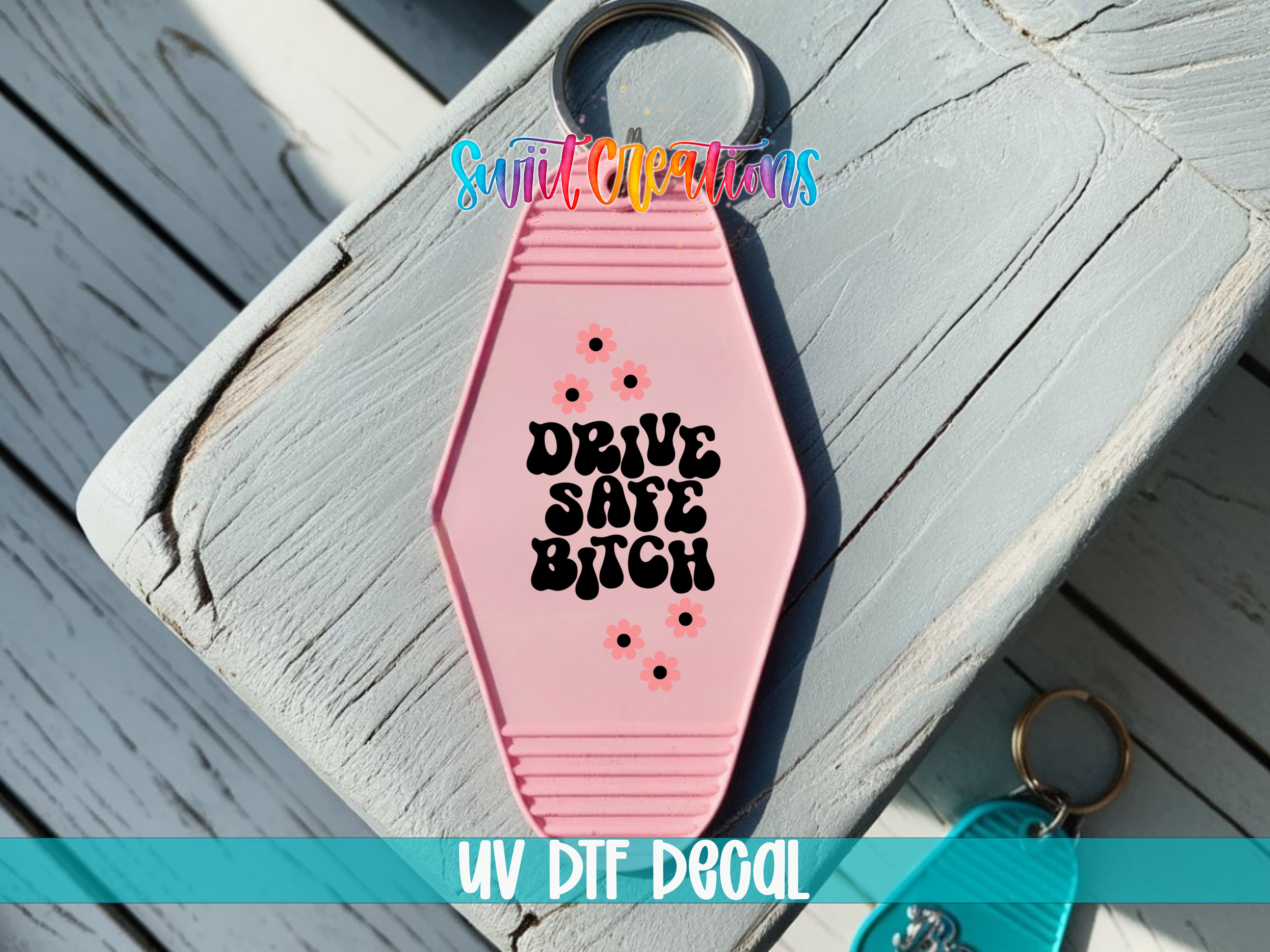 A pink keychain with the text "DRIVE SAFE BITCH" printed on it, placed on a wooden surface.
