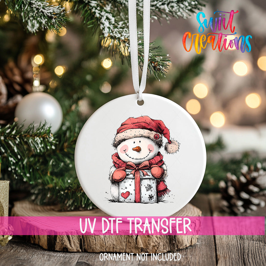 A white ornament with a snowman and a gift is hanging on a Christmas tree, with a pink ribbon around it.