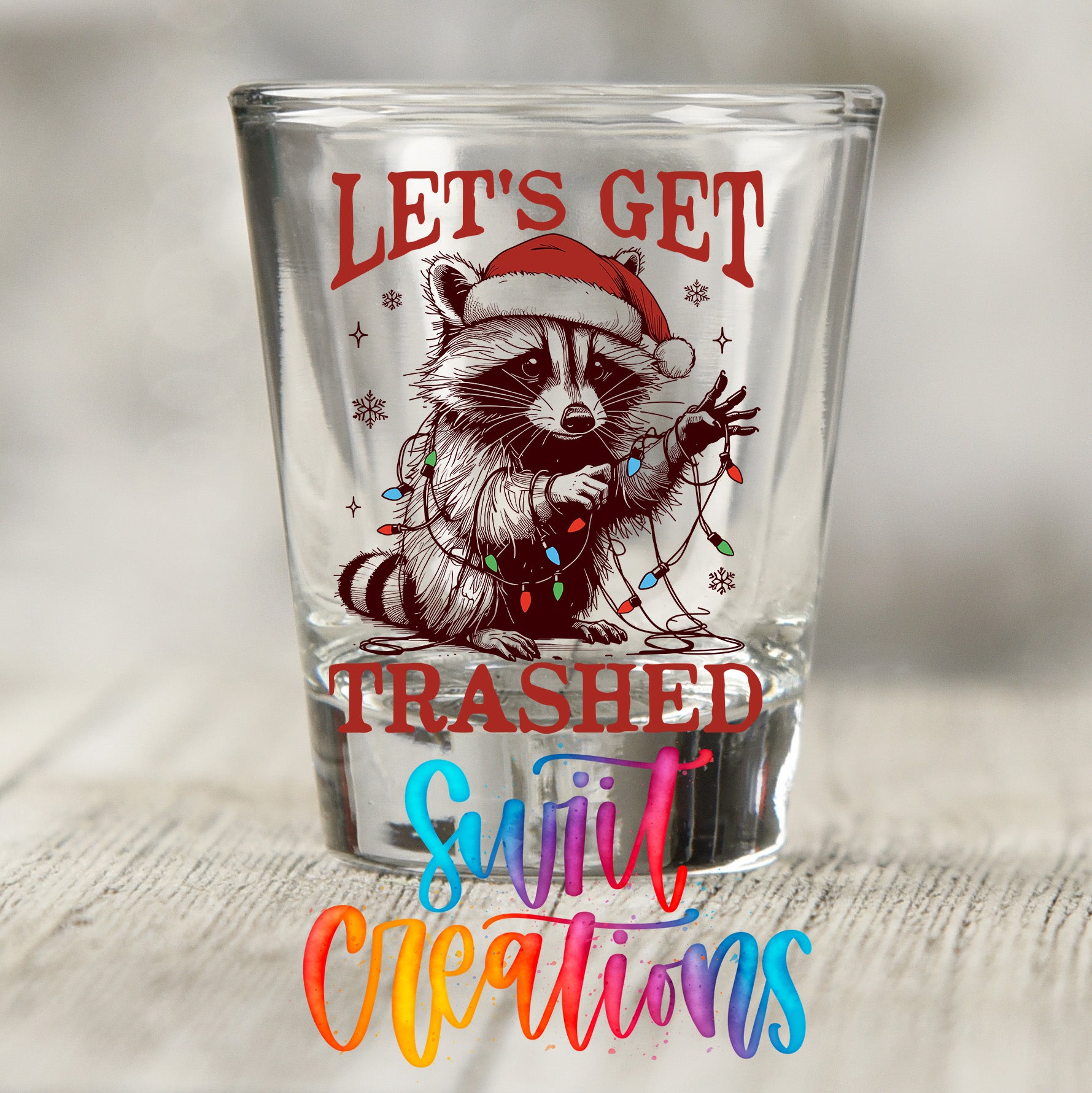 A shot glass with a raccoon wearing a Santa hat and holding Christmas lights, along with the text "Let's get trashy" and "Trashed" printed on it.