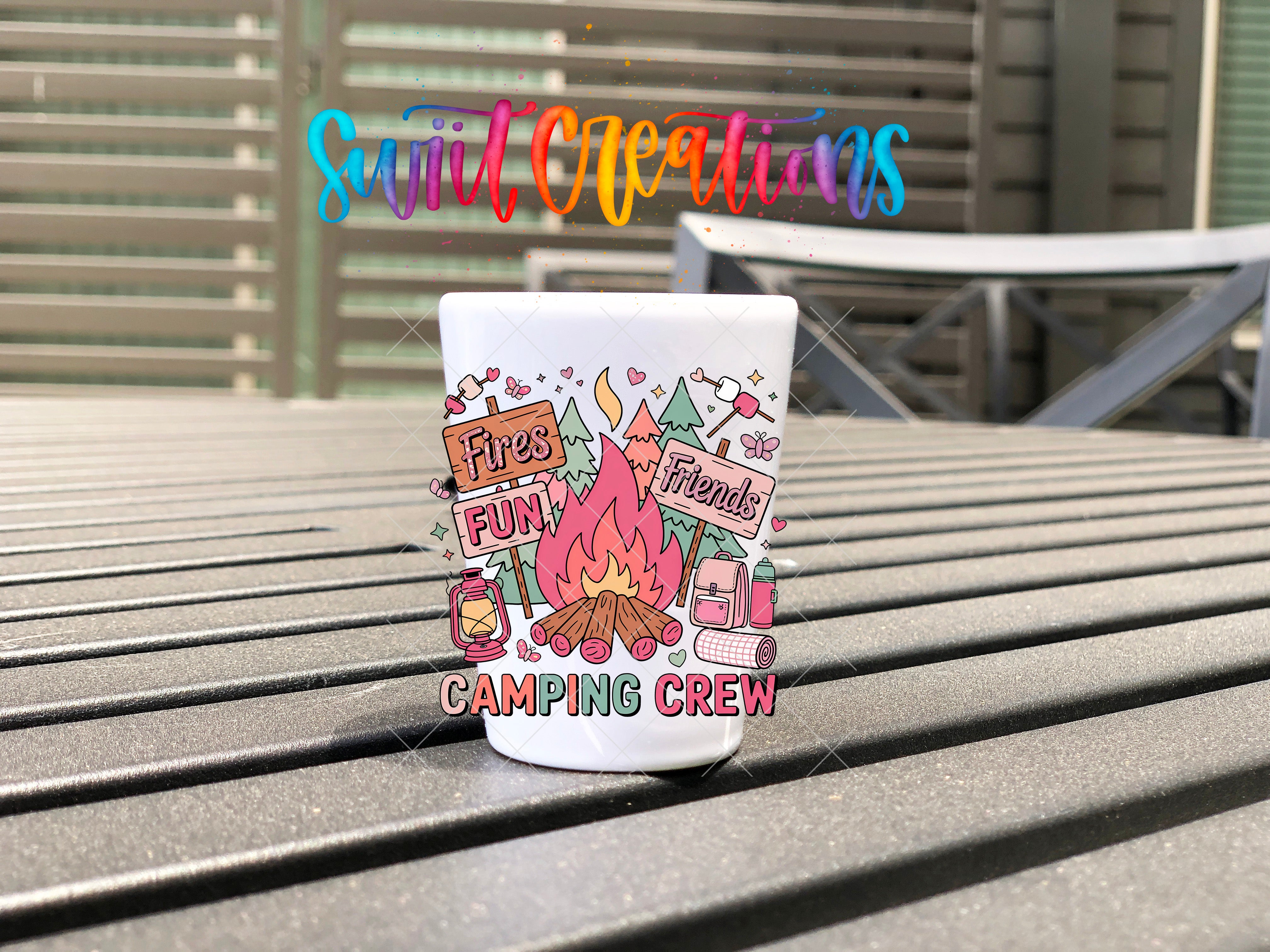 A white mug with a colorful design featuring camping-related elements, such as a campfire, tent, and camping equipment, is placed on a wooden surface.