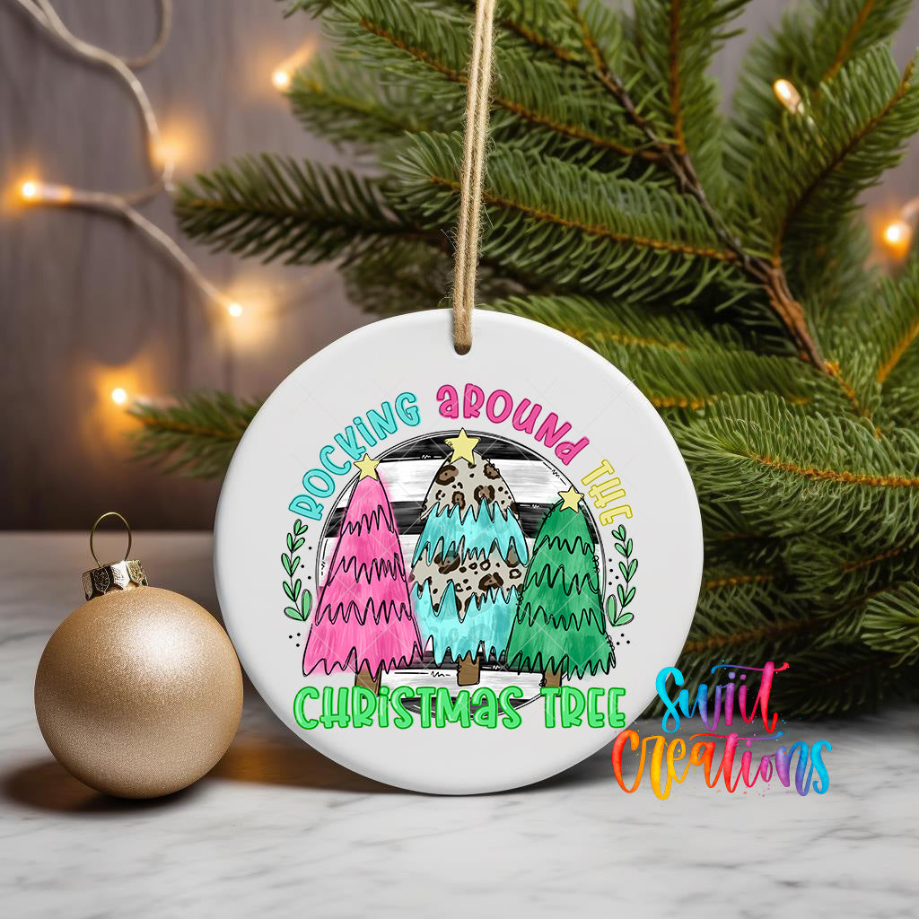 A white ceramic ornament with a colorful illustration of three Christmas trees, along with the text "Rocking Around The Christmas Tree" and "The Christmas Tree" written on it. The ornament is hanging from a string, and there is a gold ornament next to it on a table.