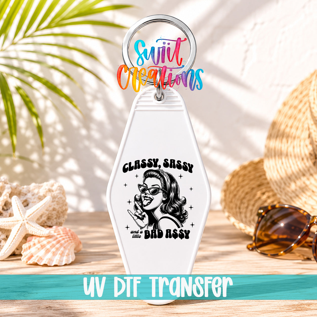 A keychain with a design featuring a woman's face and the text "Classy Sassy and Bad Ass" printed on it, along with a colorful ring around the keychain. The keychain is placed on a wooden surface, with sunglasses and a straw hat visible in the background.