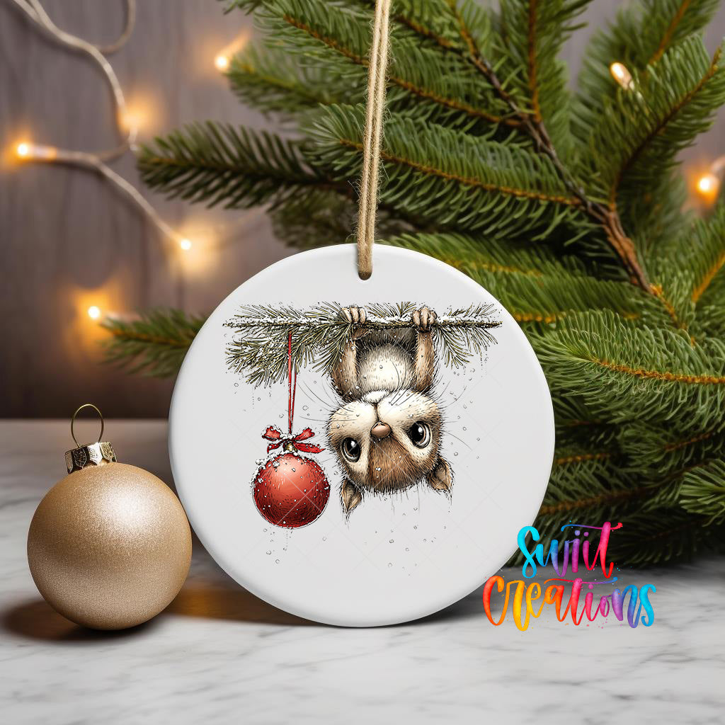 A white ceramic ornament with a playful illustration of a cat hanging from a branch, holding a red ornament, and surrounded by a Christmas tree and twinkling lights.