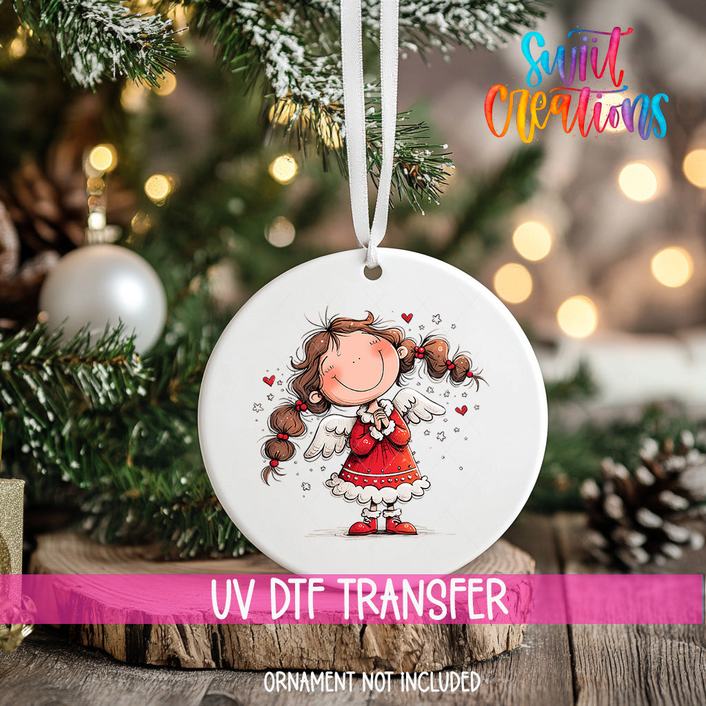 A white ceramic ornament with a smiling girl with long hair and wings, surrounded by stars, is hanging on a Christmas tree.