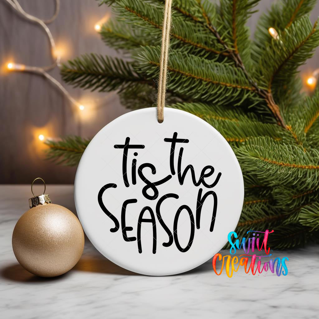 A white ornament with the text "Tis the Season" written on it, hanging from a string, is placed next to a gold ornament on a Christmas tree.
