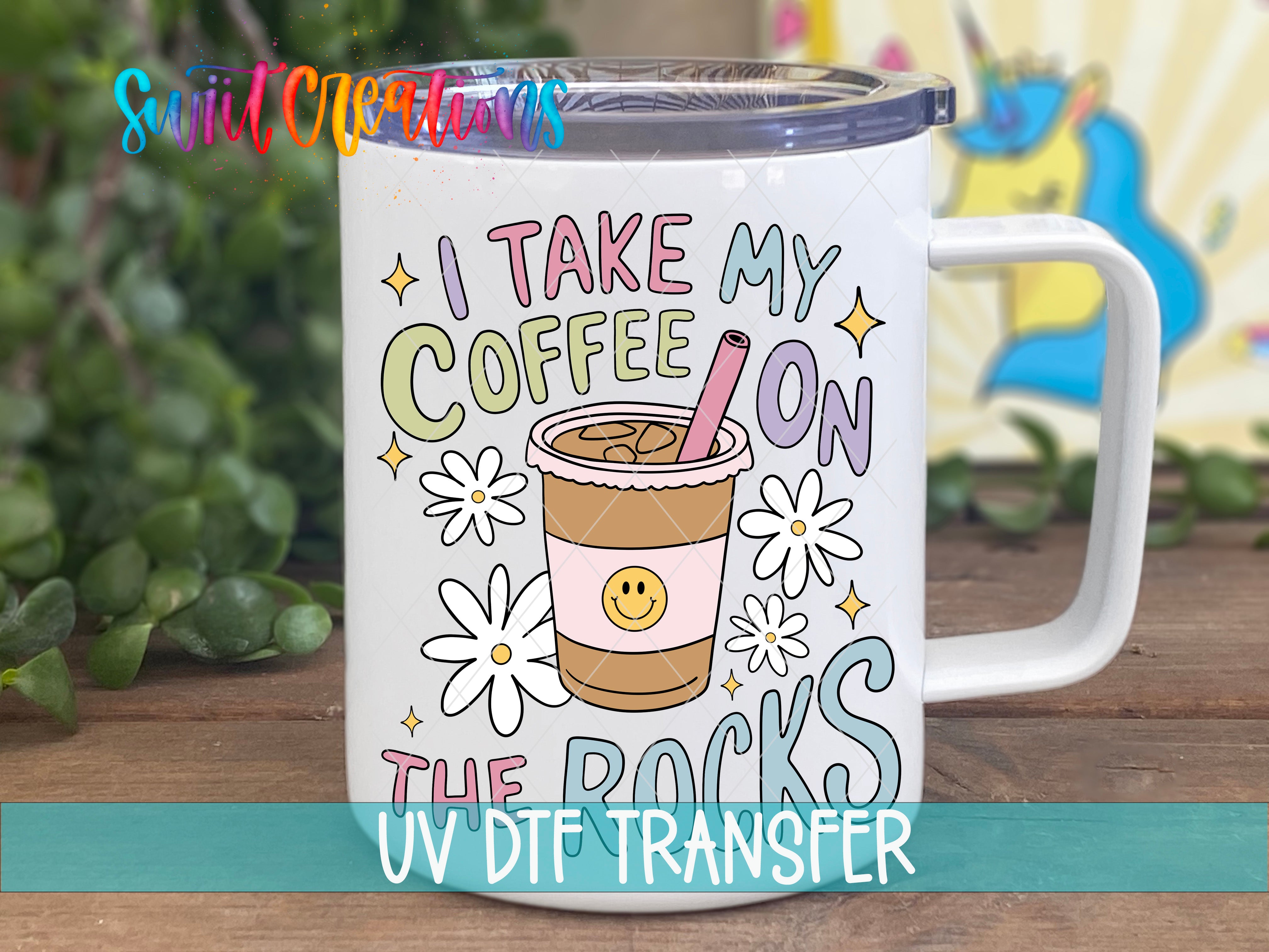 A white mug with a colorful design featuring a coffee cup and flowers, along with the text "I TAKE MY COFFEE ON THE ROCKS" and "UV Dtf Transfer".