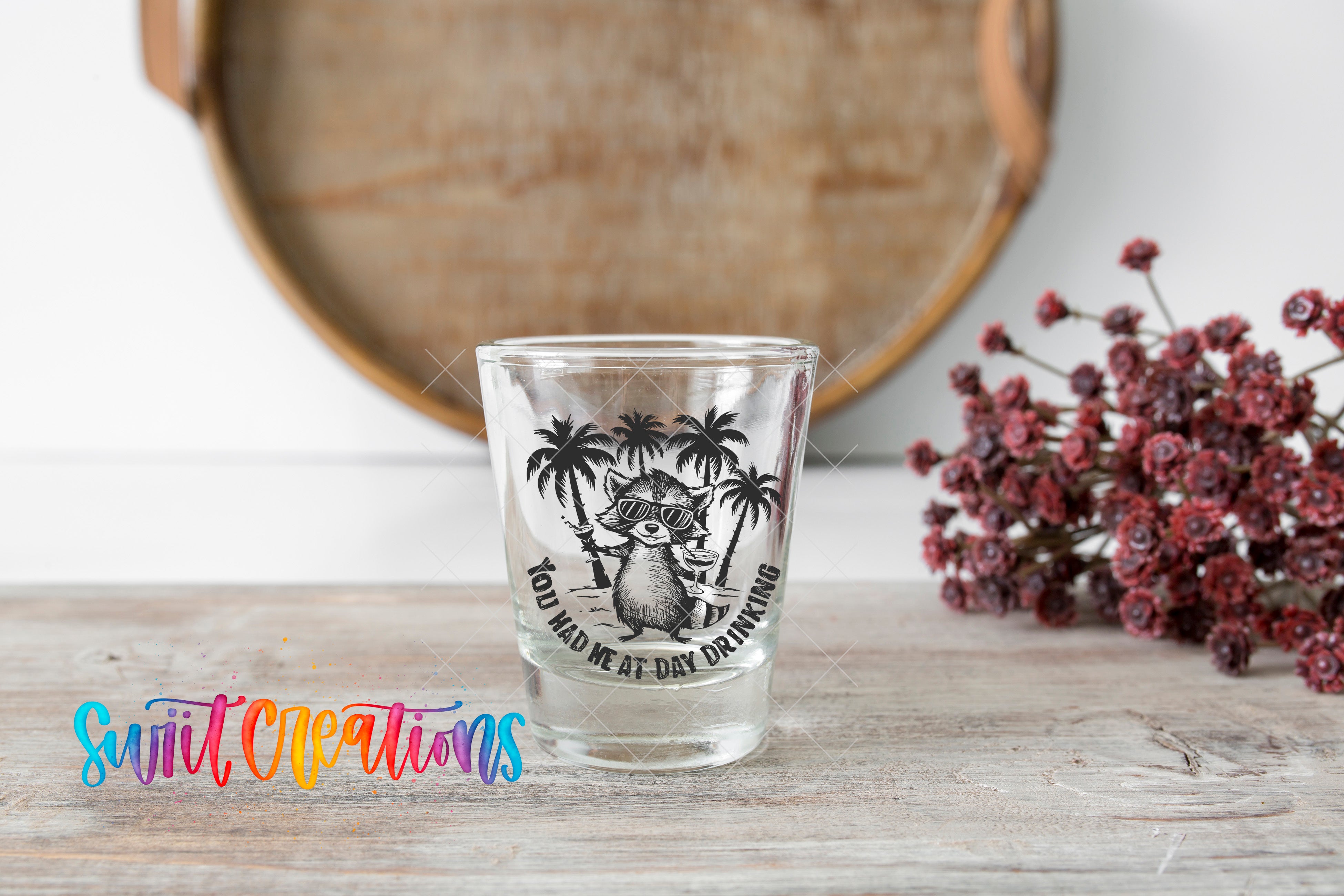 A shot glass with a raccoon design and the text "You had one at day drinking" is placed on a wooden surface, with a bunch of red flowers in the background.