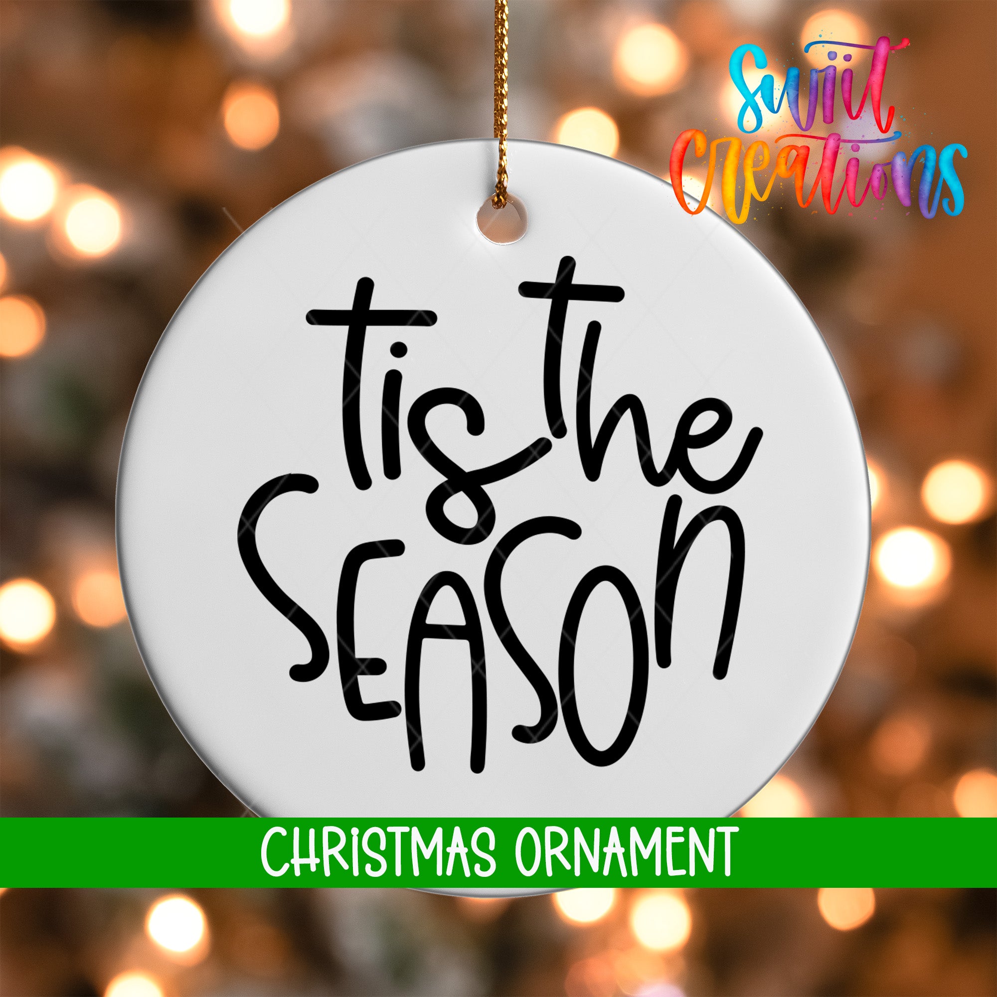 A white Christmas ornament with the text "Tis the SEASON" written on it, hanging against a background of colorful, blurred lights.