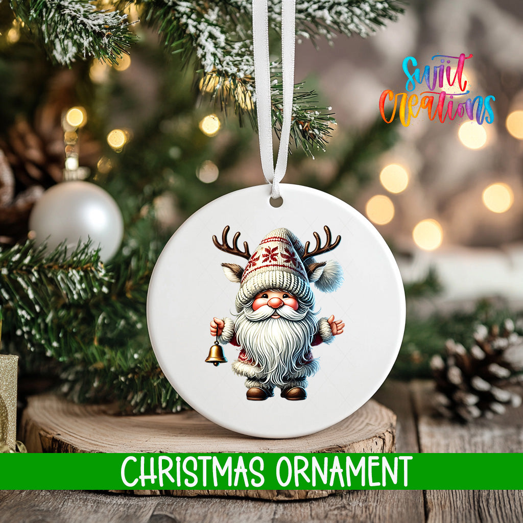 A white Christmas ornament featuring a gnome with antlers and a bell, hanging on a tree branch with a blurred background of lights and greenery.
