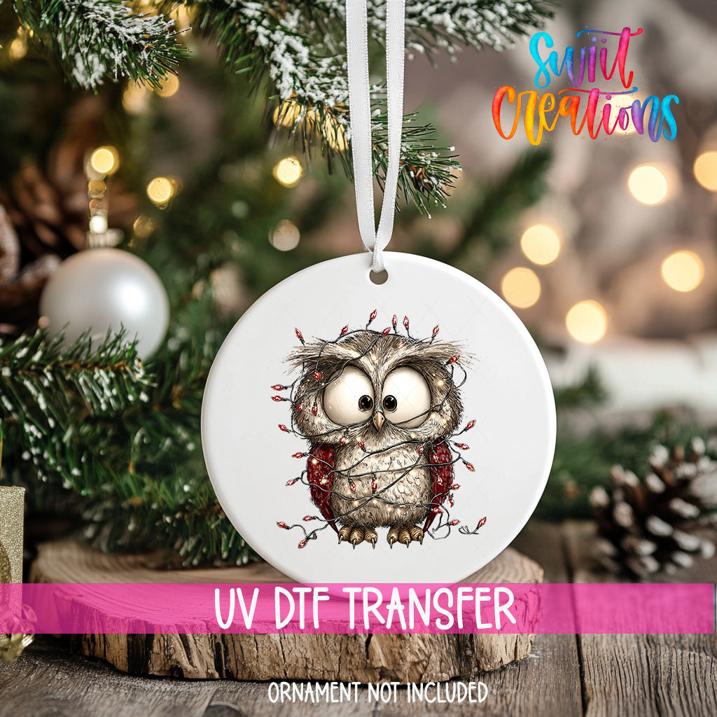 A white ornament with a cartoon owl design is hanging on a Christmas tree, with a blurred background of lights and pine branches.