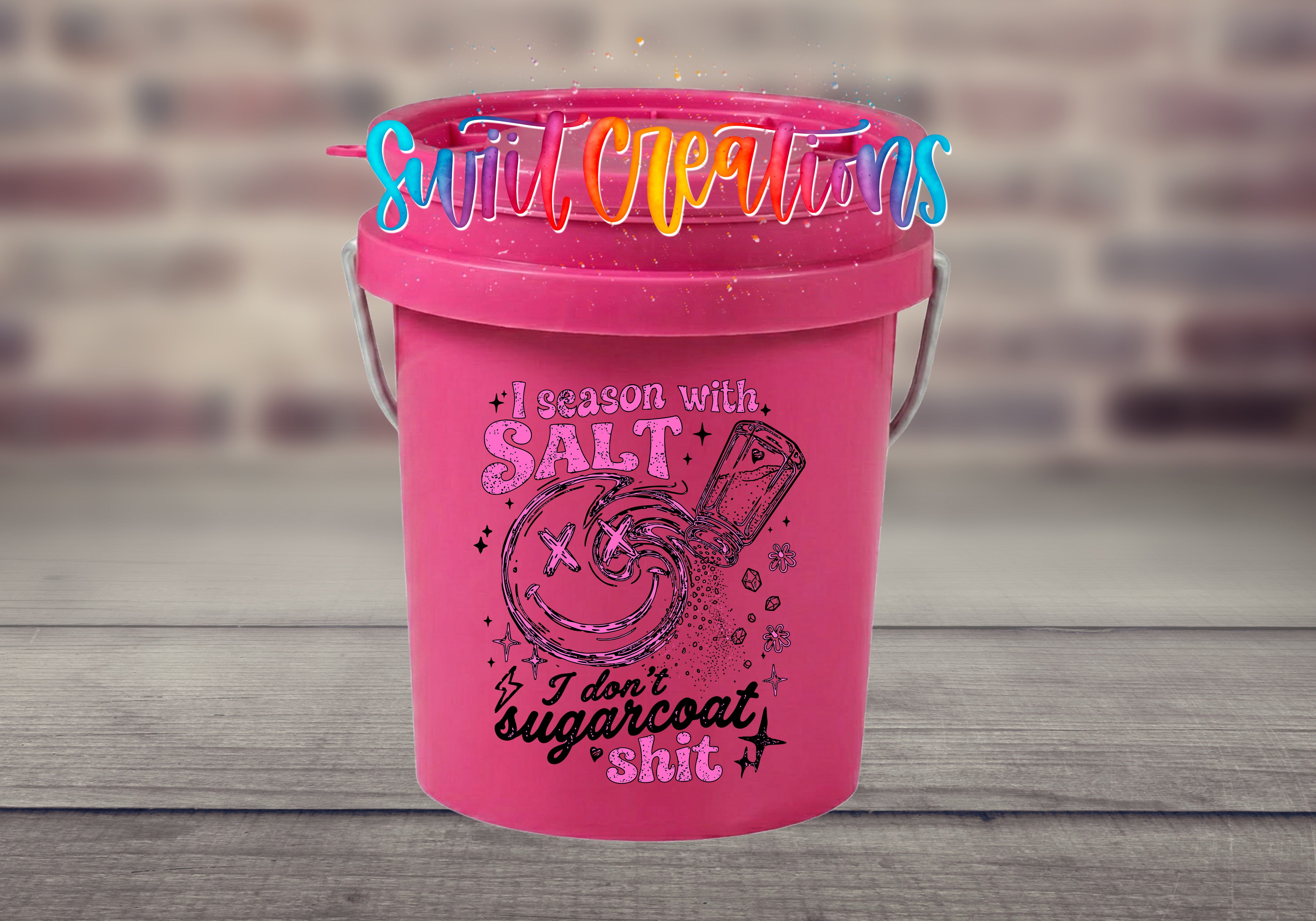 A pink bucket with a humorous text and graphics printed on it, placed on a wooden surface against a blurred brick wall background.