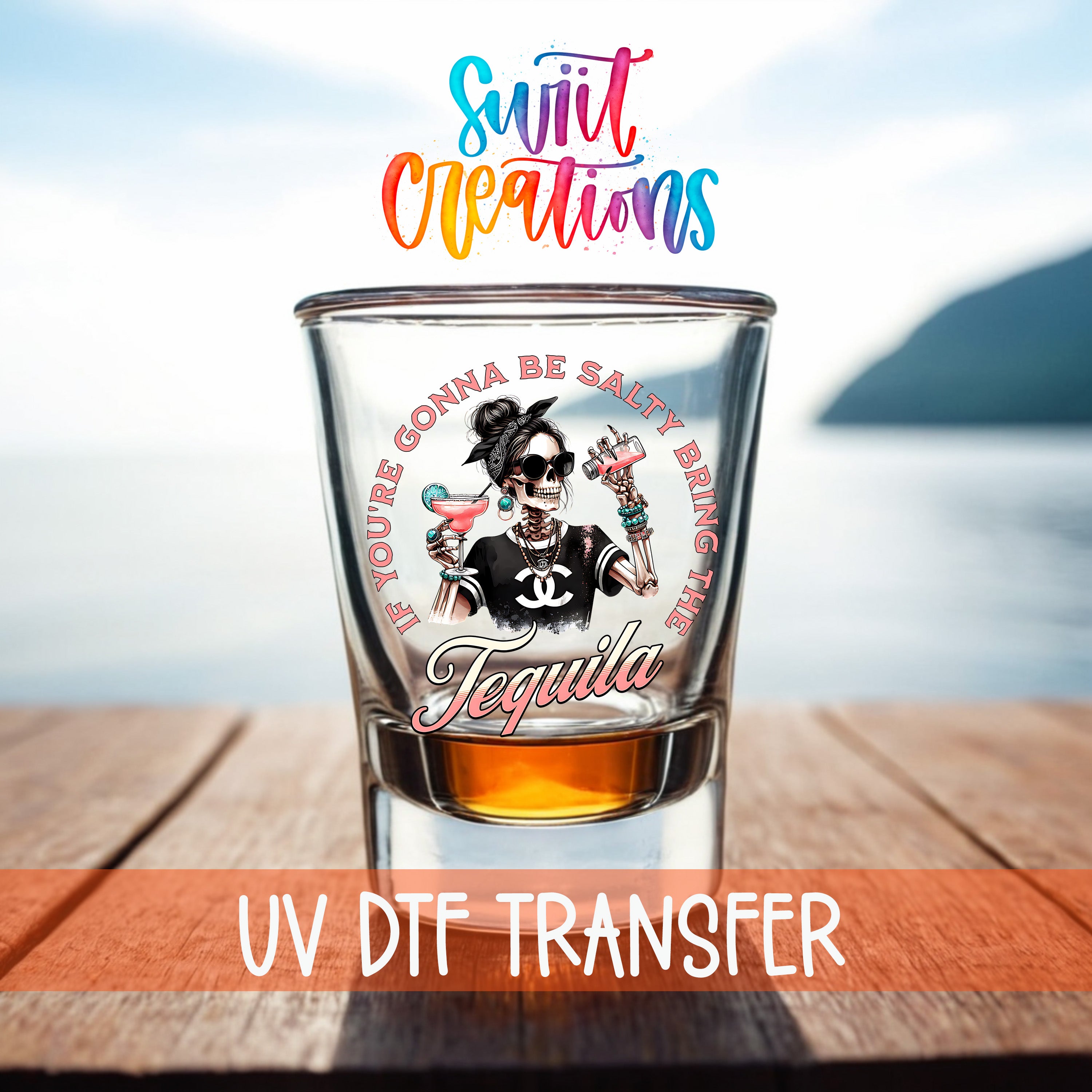 A shot glass with a colorful design featuring a woman with a skull-like face and the text "If you're gonna be salty bring the tequila" printed on it, placed on a wooden surface with a body of water in the background.