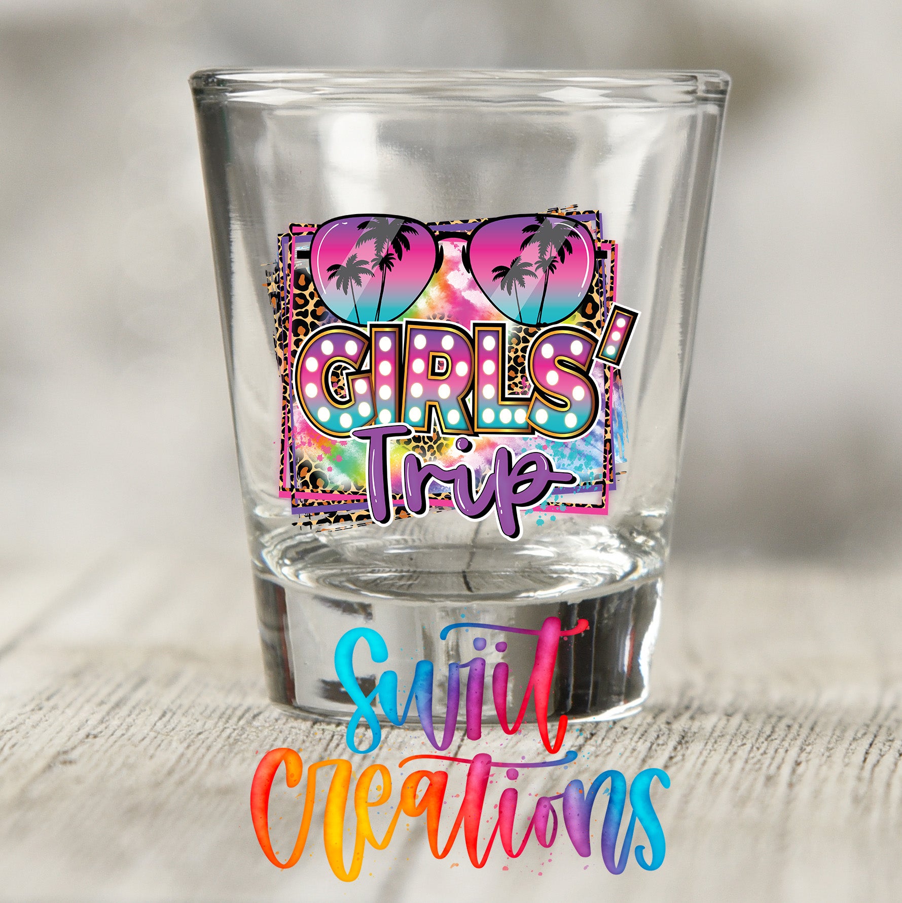 A clear shot glass with a colorful graphic design featuring sunglasses and palm trees, along with the text "GIRLS' TRIP" and "Sweet Creations" written below it.