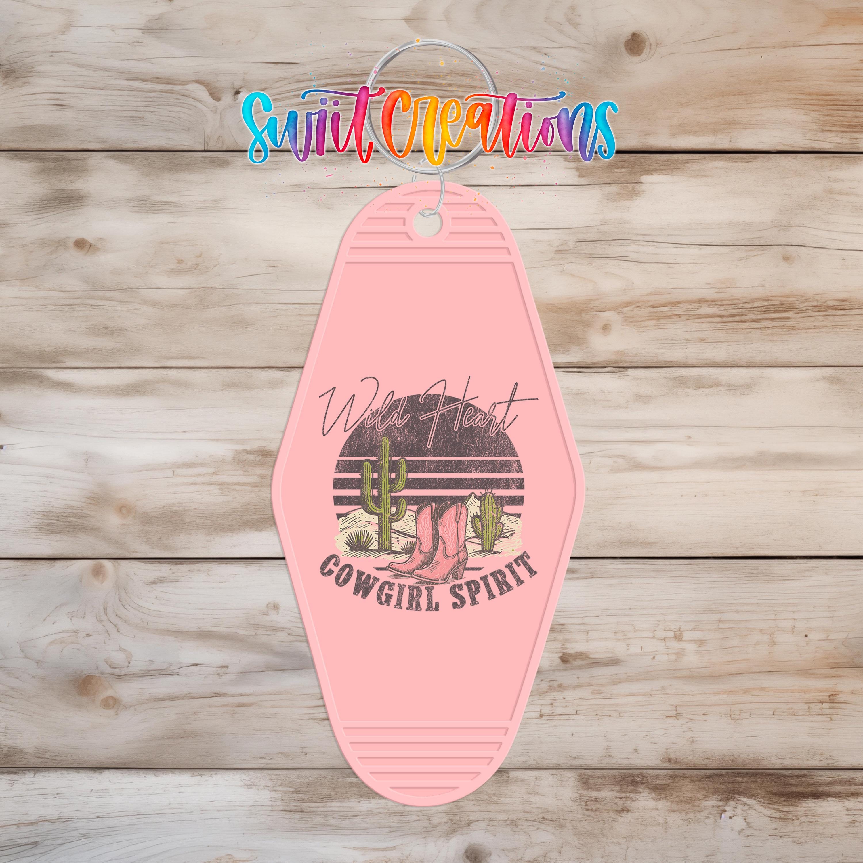 A pink keychain with a graphic design featuring a cactus and the text "Wild Heart Cowgirl Spirit" printed on it, hanging on a wooden surface.