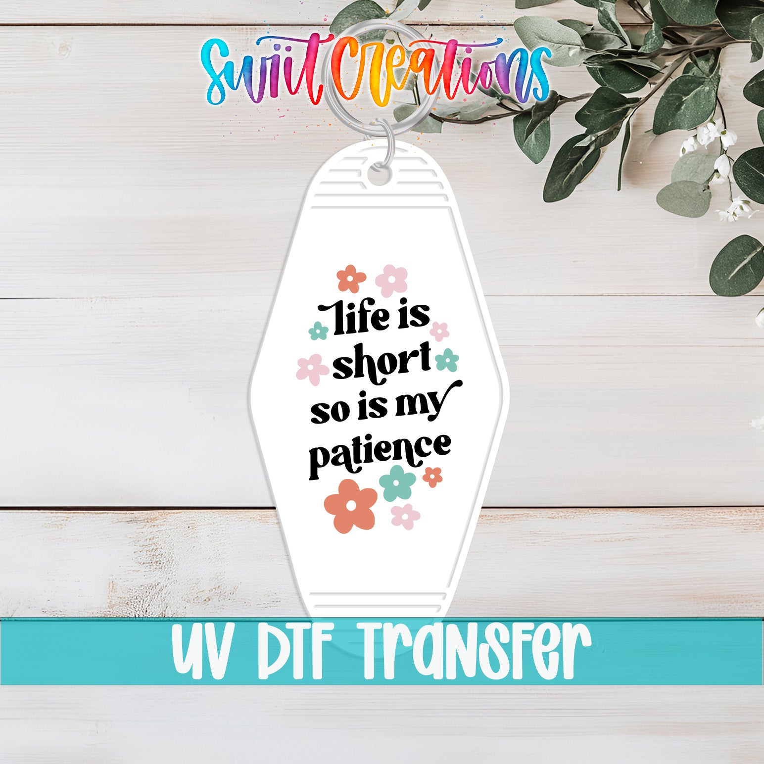 A keychain with the text "Life is short so is my patience" printed on it, along with a floral design. The keychain is placed on a wooden surface, and there is a plant in the background.