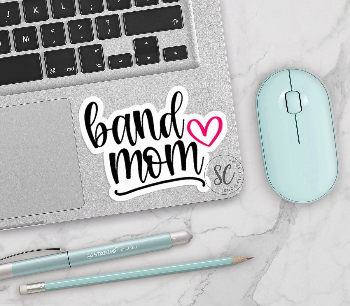 Pink Heart Band Mom vinyl sticker on a silver laptop next to a mint mouse and pens