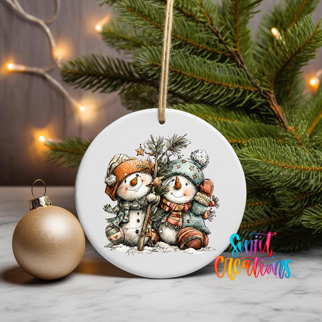 A white ceramic ornament featuring two snowmen is displayed on a table, with a gold ornament and a Christmas tree in the background.