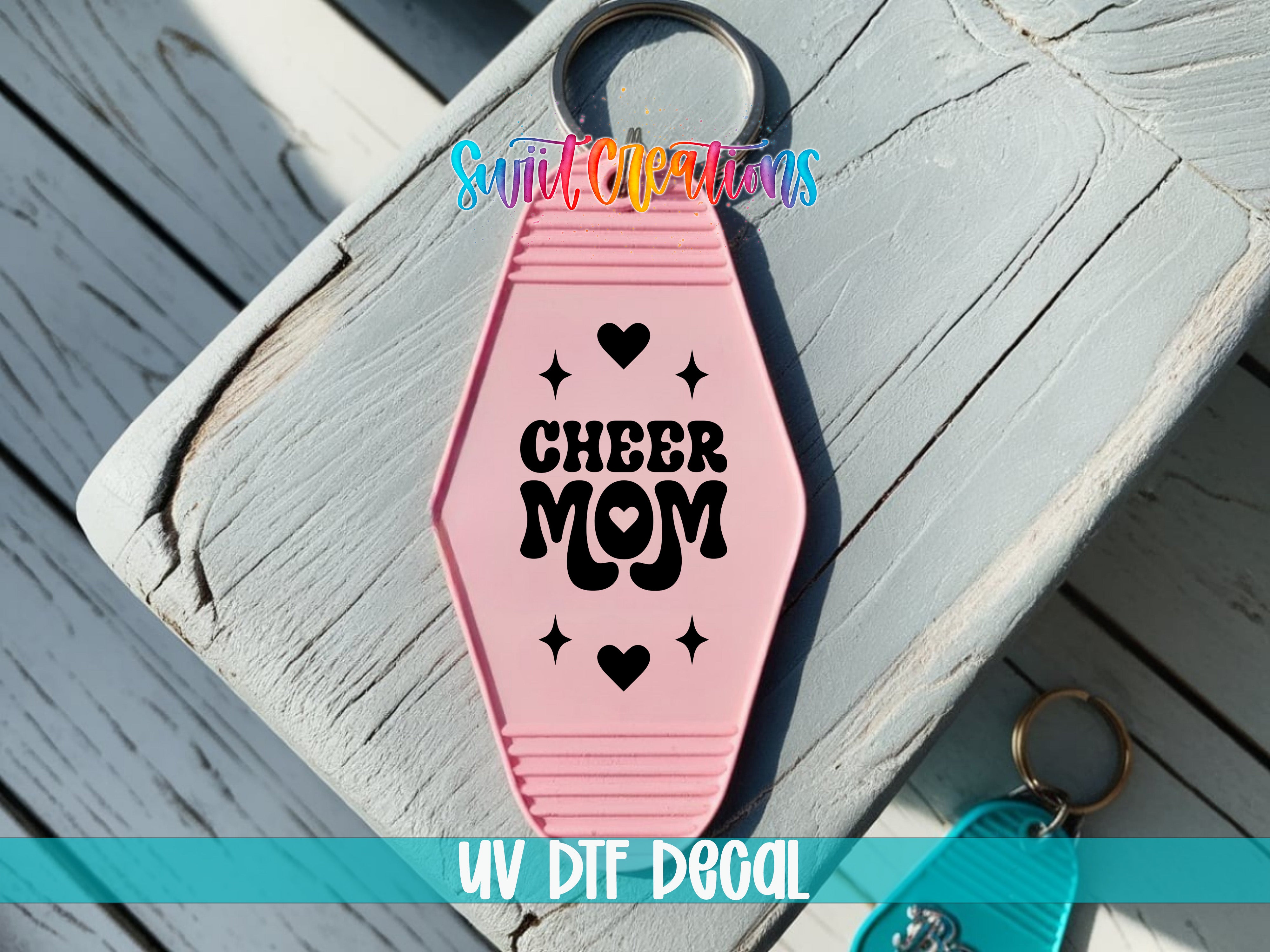 A pink keychain with the text "CHEER MOM" printed on it, placed on a wooden surface.