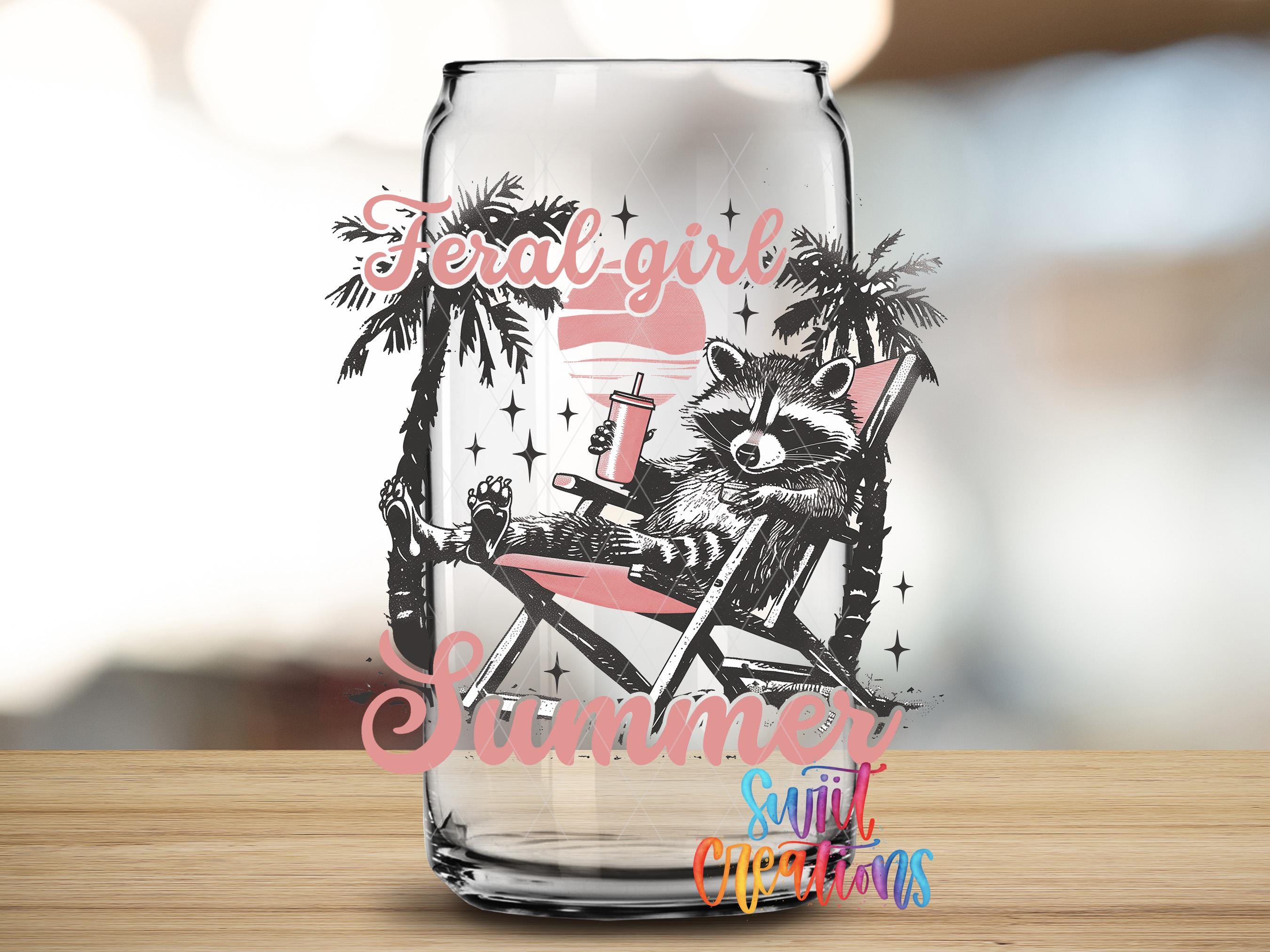 A glass can with a raccoon sitting in a beach chair, holding a drink, and surrounded by palm trees and stars, with the text "Feral Girl Summer" and "Sunlit Creations" printed on it.
