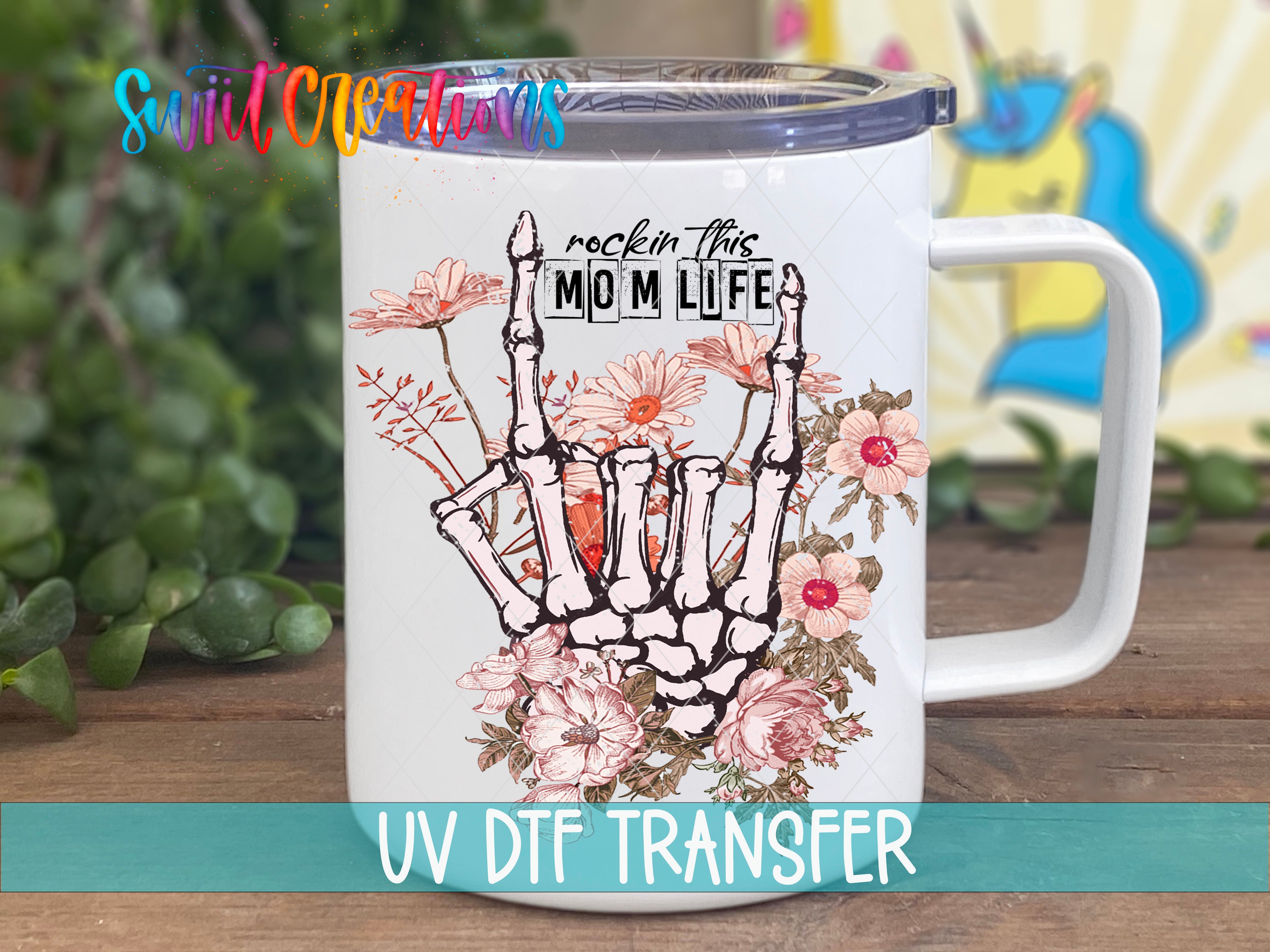 A white ceramic mug with a floral design and a skeleton hand gesture, along with the text "rock in this mom life" printed on it.