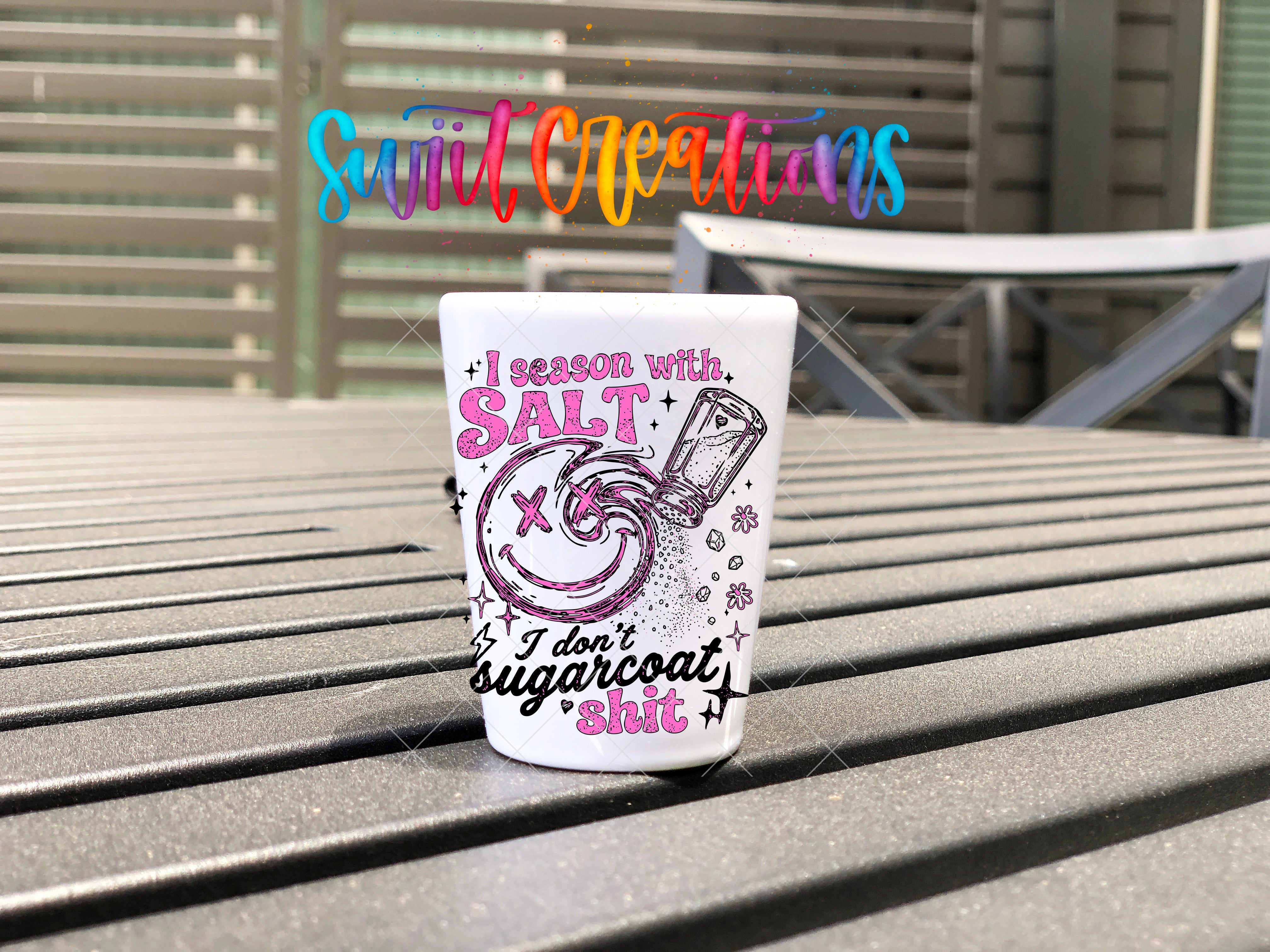 A white shot glass with a colorful design and text on it, placed on a wooden surface.