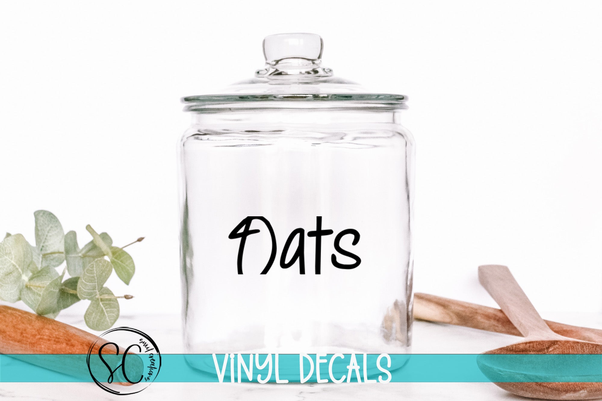 A clear glass jar with the word "Oats" written on it, placed on a white surface next to some wooden utensils.