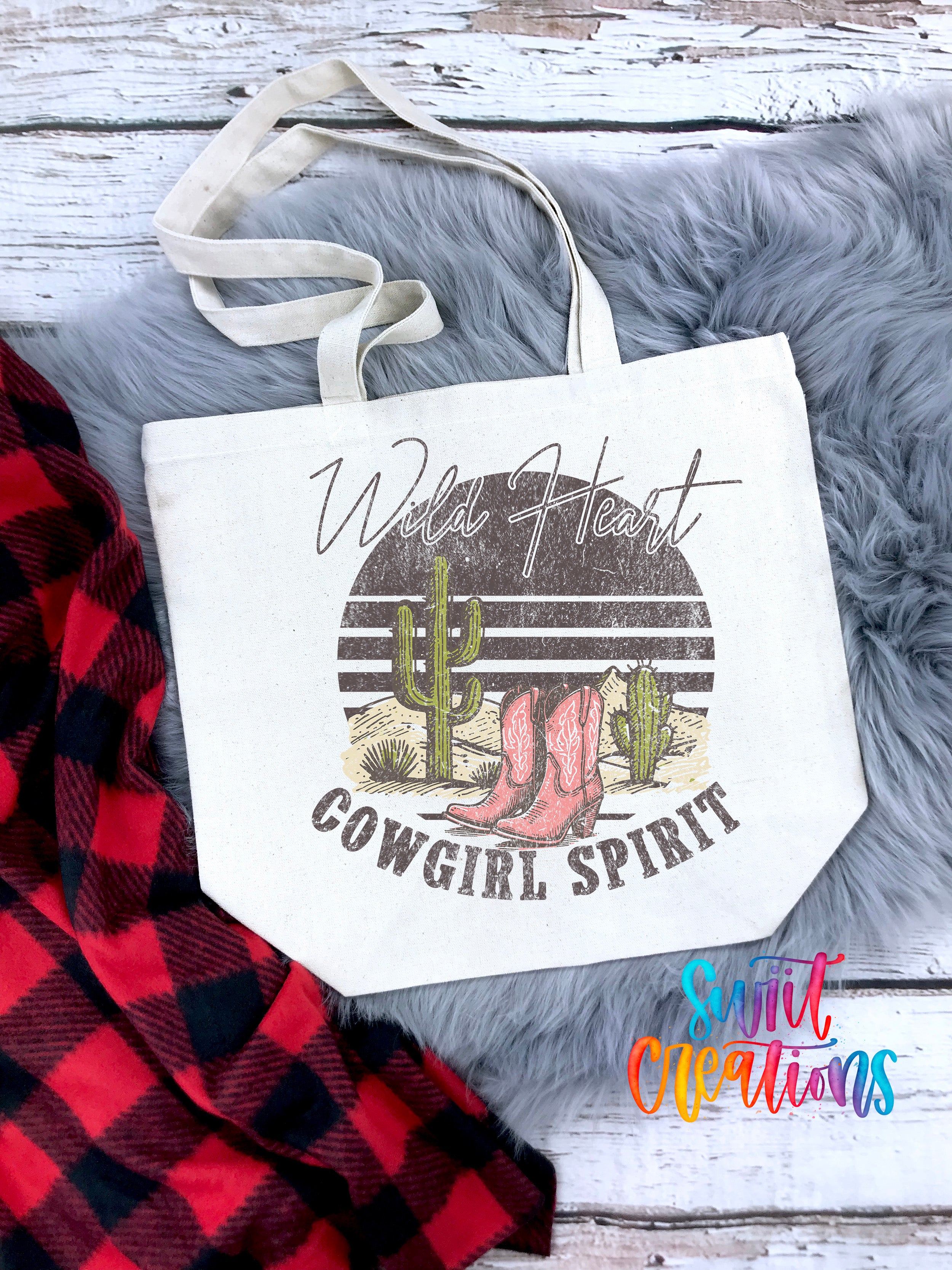 A white tote bag with a graphic design featuring a cactus and cowboy boots, along with the text "Wild Heart Cowgirl Spirit". The bag is placed on a gray furry surface, and there is a red and black plaid blanket nearby.