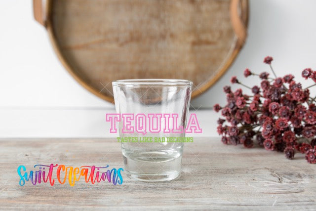 A clear shot glass sits on a wooden surface, with a wooden tray and a bouquet of red flowers in the background.