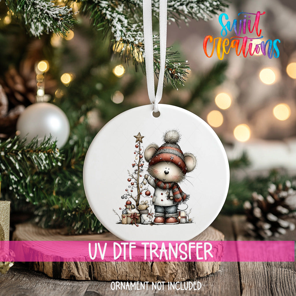 A white ornament with a cartoon mouse and Christmas tree is hanging on a tree, with a pink ribbon around it.