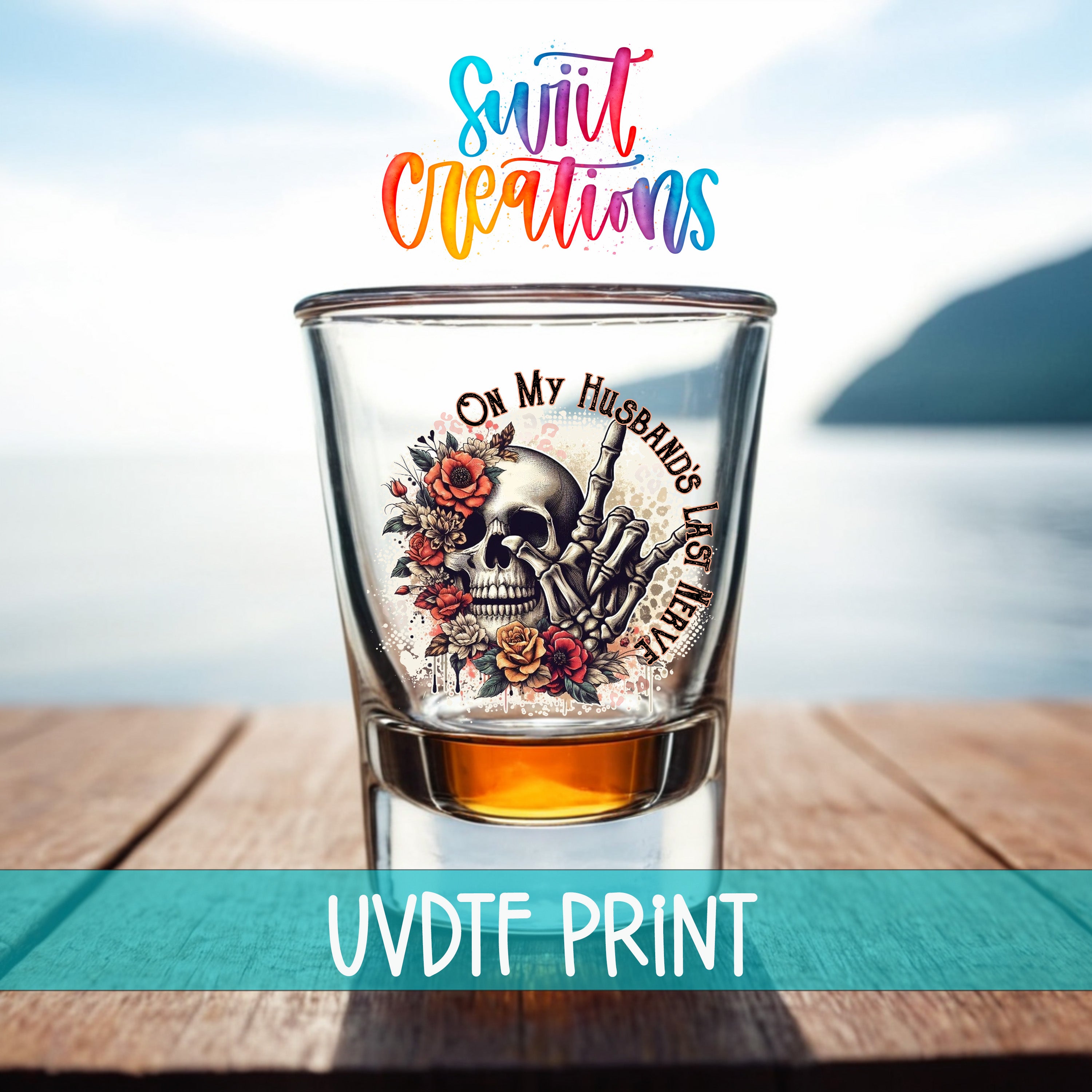 A shot glass with a skull design and floral elements, placed on a wooden surface with a body of water in the background.