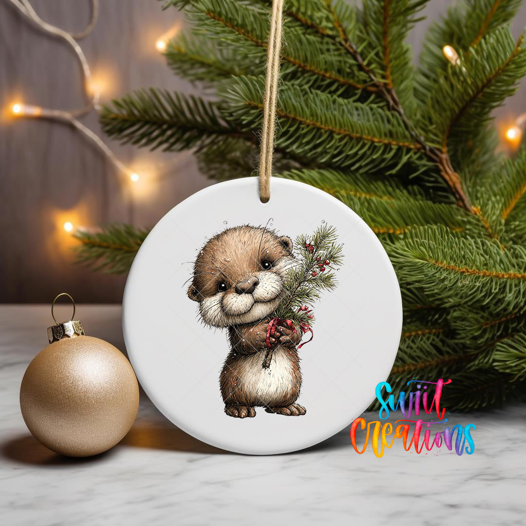 A ceramic ornament featuring a cute otter holding a pine branch, placed on a marble surface next to a gold ornament and a Christmas tree with twinkling lights.